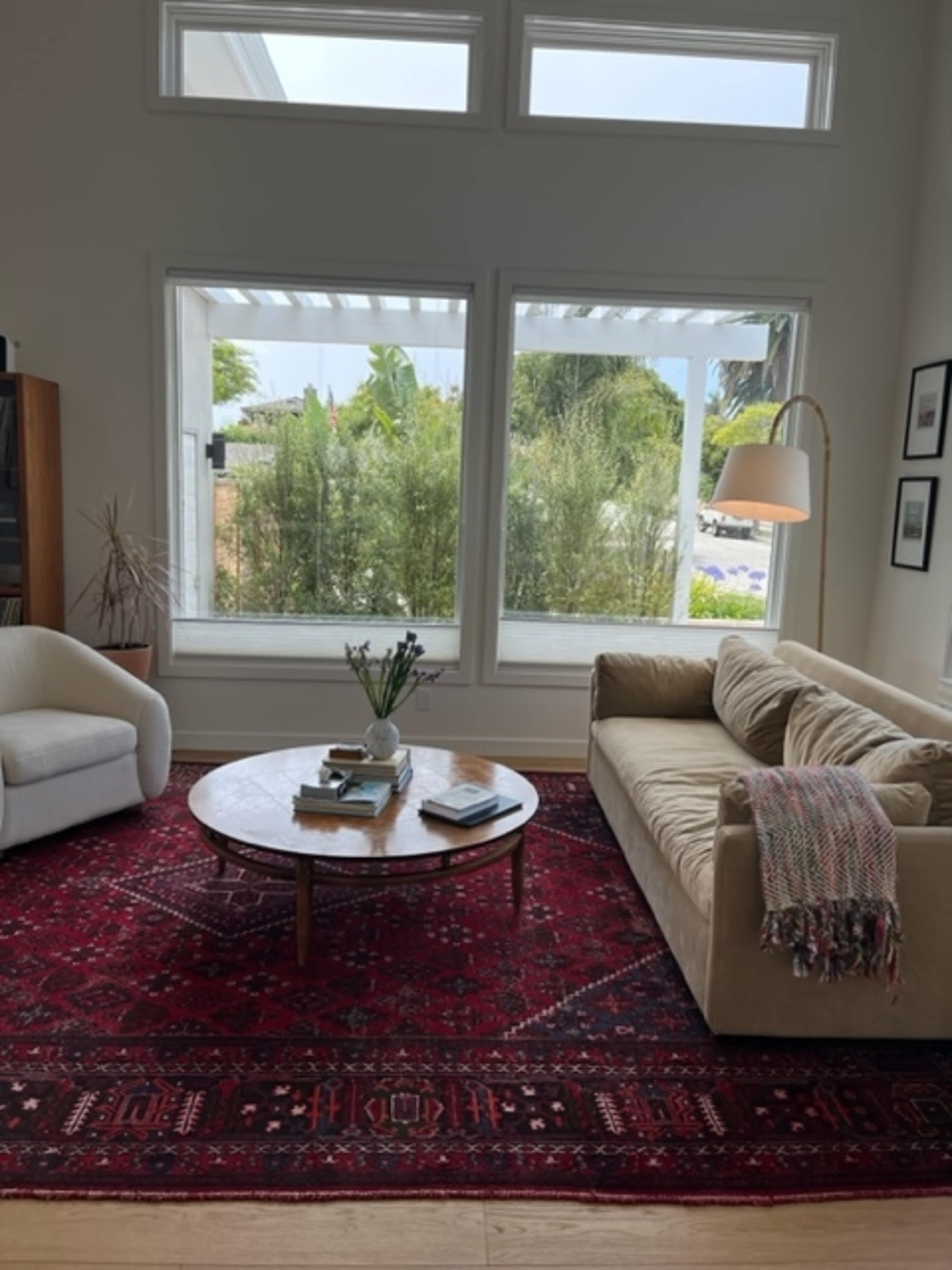A living room with a light-colored sofa and armchair arranged around a round coffee table on a red patterned rug, with large windows offering a view of greenery outside.