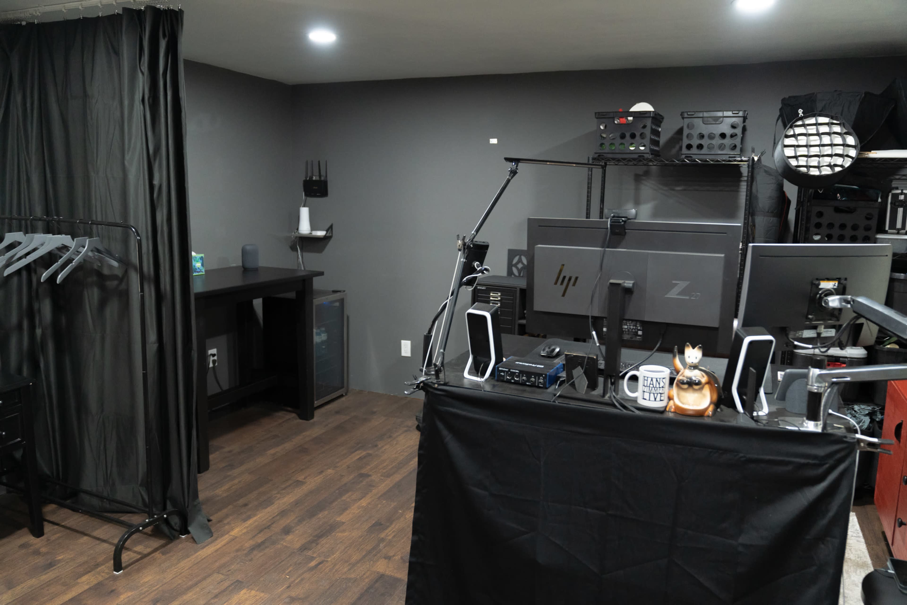 The image shows a workspace featuring a computer setup on a shelf, a black curtain, and a table with hangers against a gray wall.
