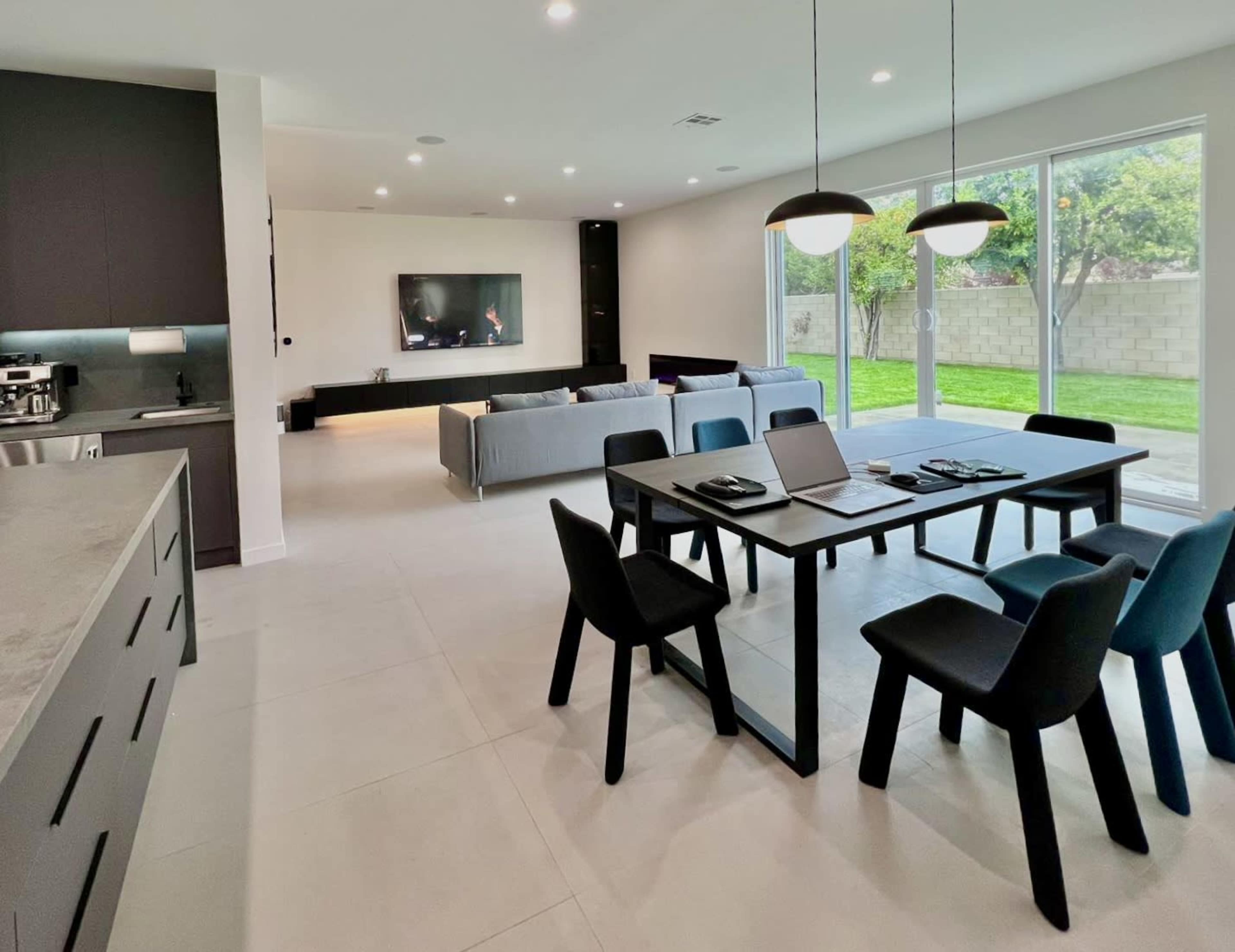 A spacious, modern living area with a dining table, black chairs, a couch, and large windows overlooking a grassy yard.