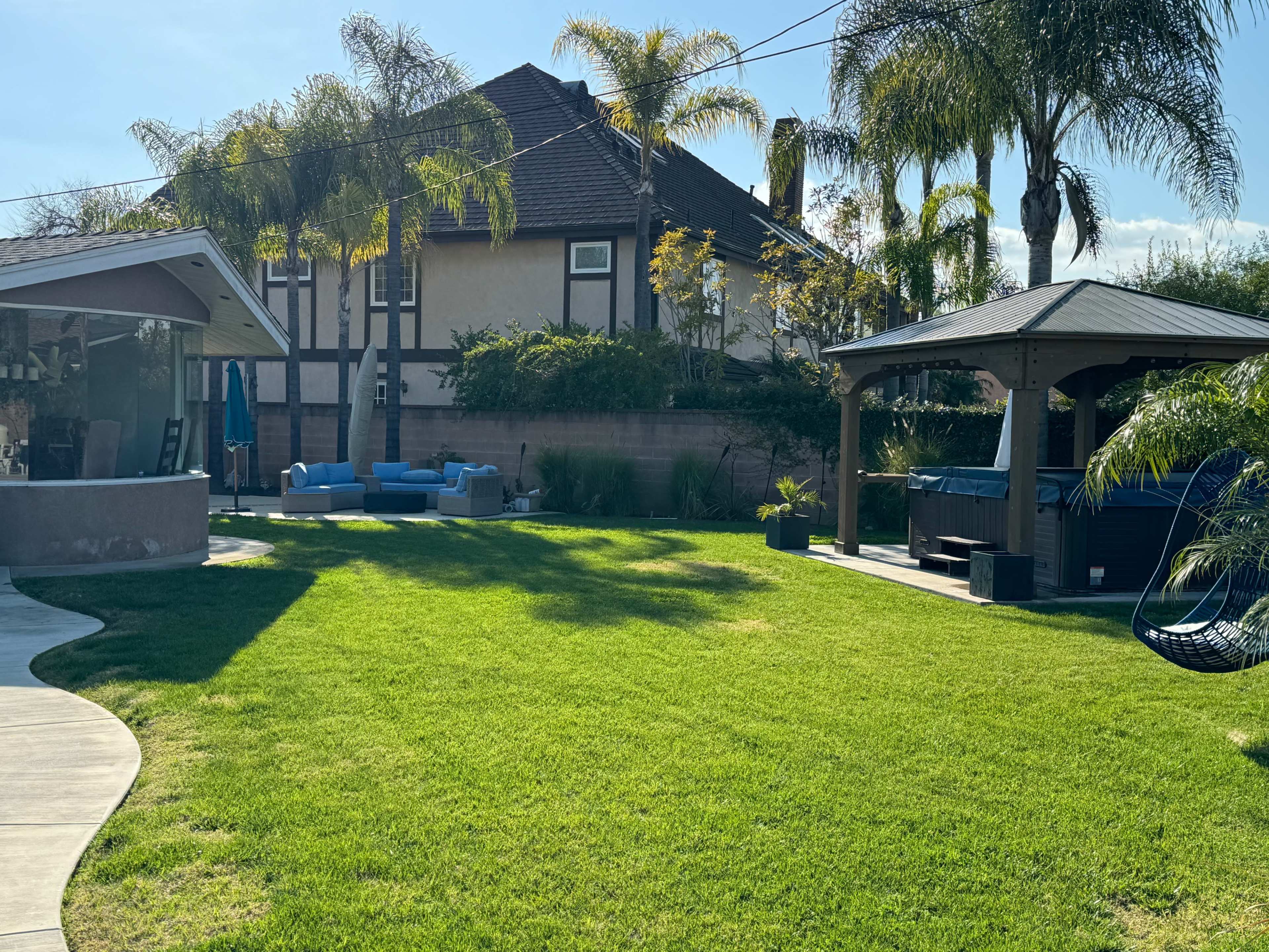 A well-maintained backyard featuring a spacious lawn, a gazebo, a hot tub, and palm trees, with a house in the background.