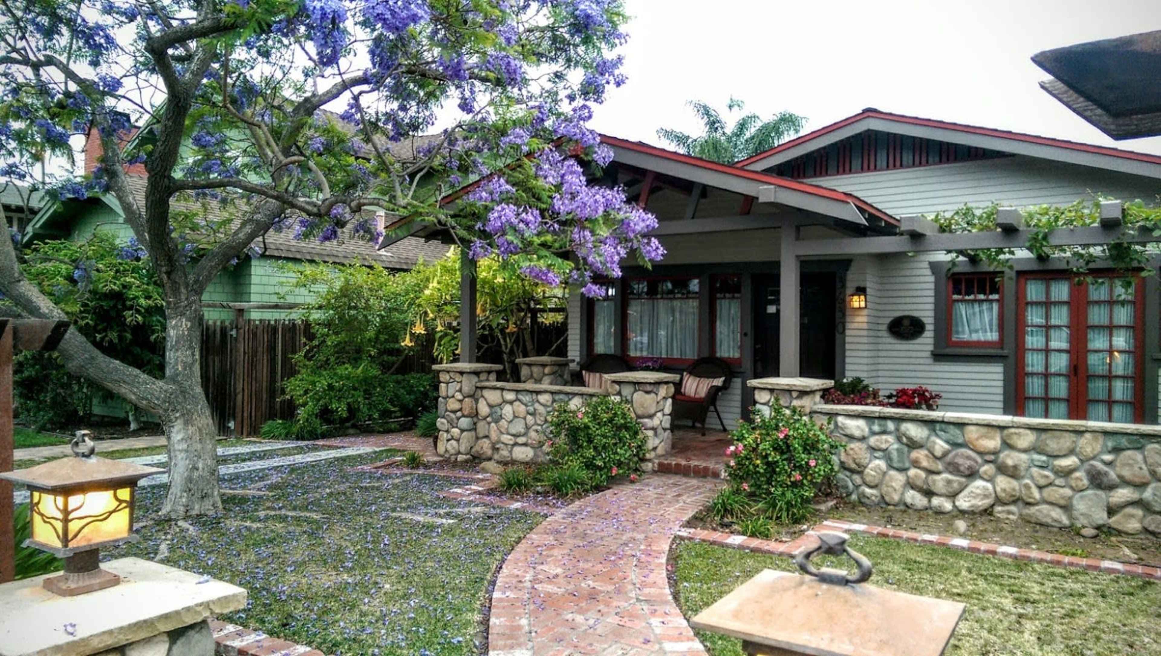 A charming bungalow features a stone pathway leading to its front porch, surrounded by a landscaped yard with purple flowering trees.