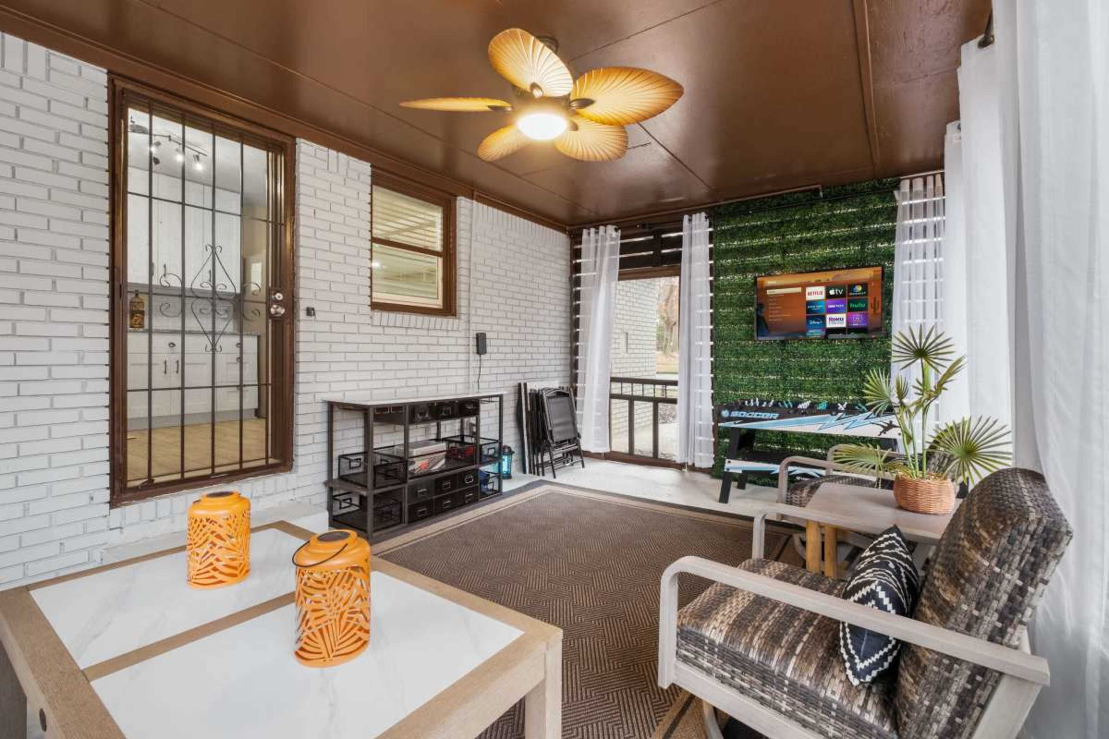 The image shows a cozy sunroom with white brick walls, a ceiling fan, a TV on the wall, and furniture arranged around a table with decorative lanterns.