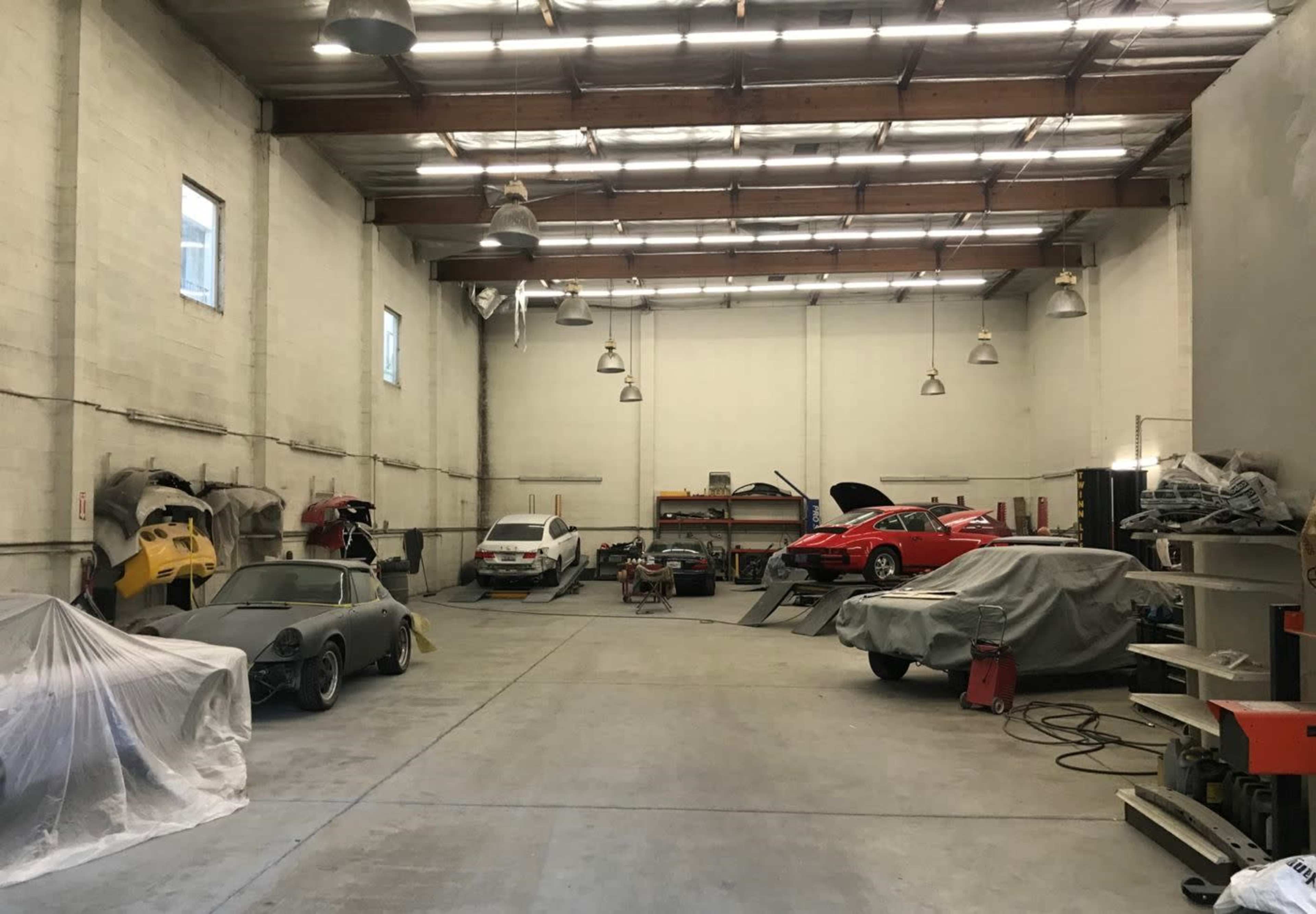 A car workshop features several vehicles, some covered and others being worked on, within a spacious industrial building.