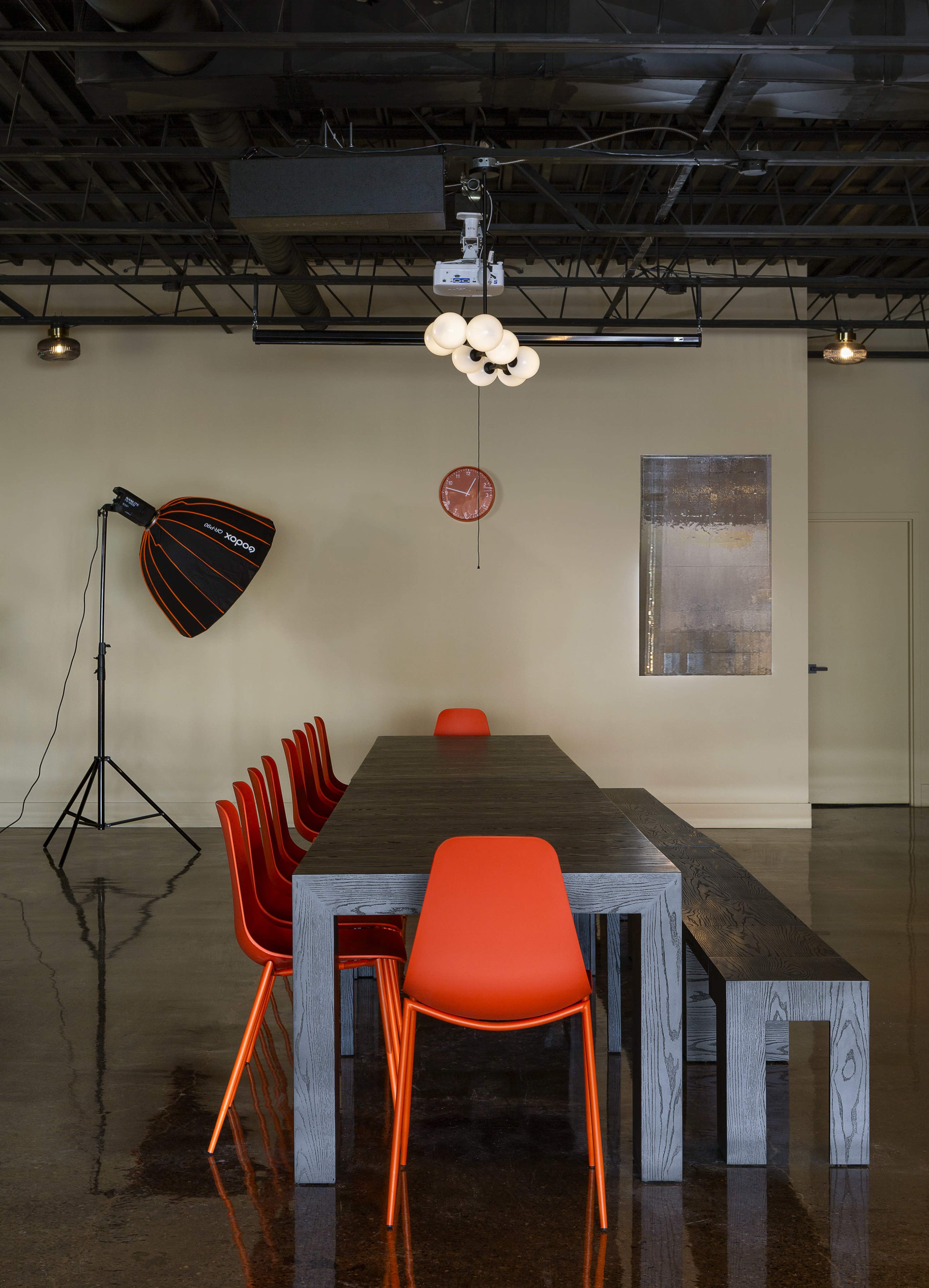 A long, rectangular wooden table with orange chairs is positioned in a modern conference room featuring industrial lighting and decor.