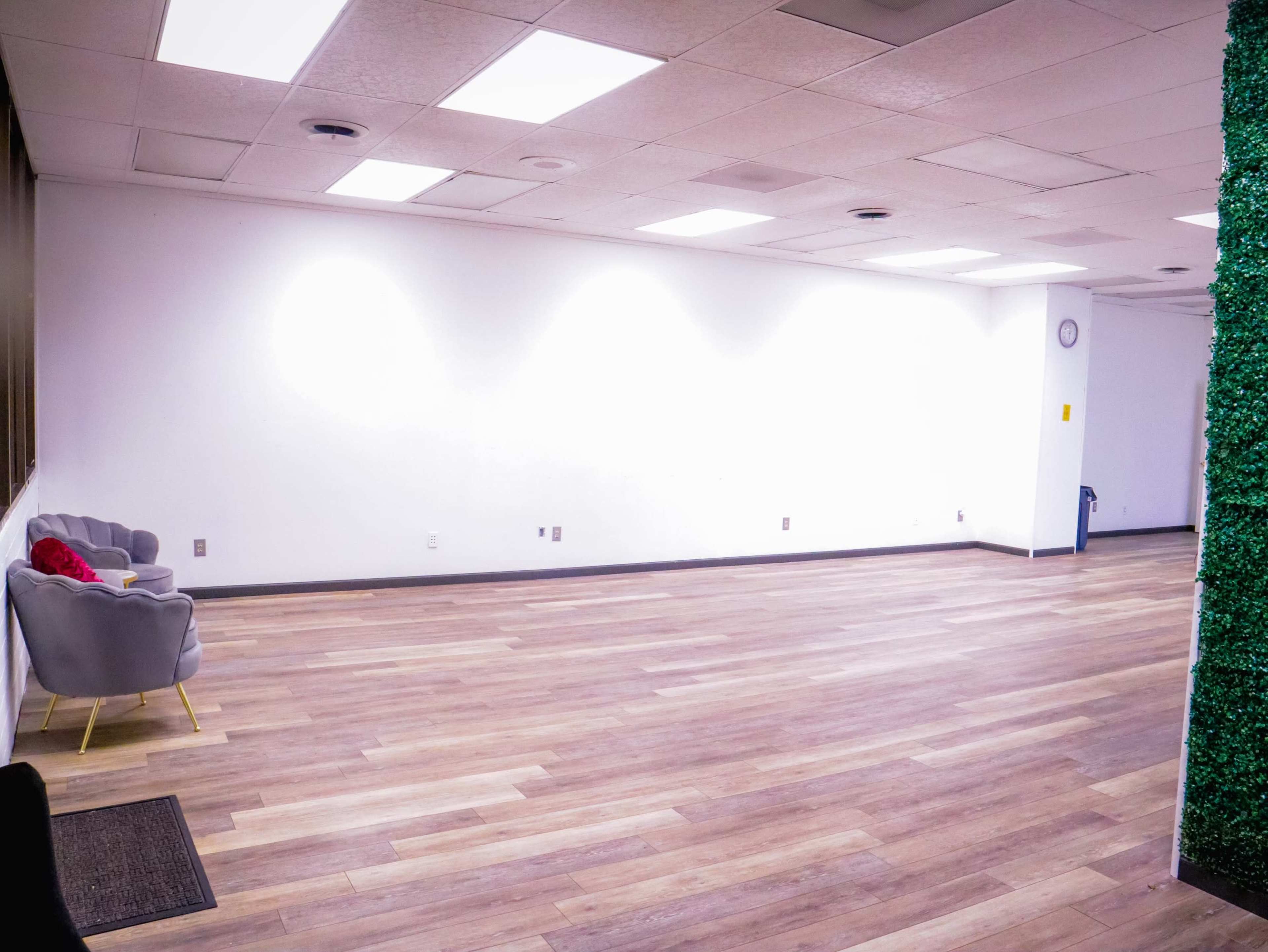 An empty room with hardwood floors, featuring a gray armchair and a small area covered in green plants on one side.