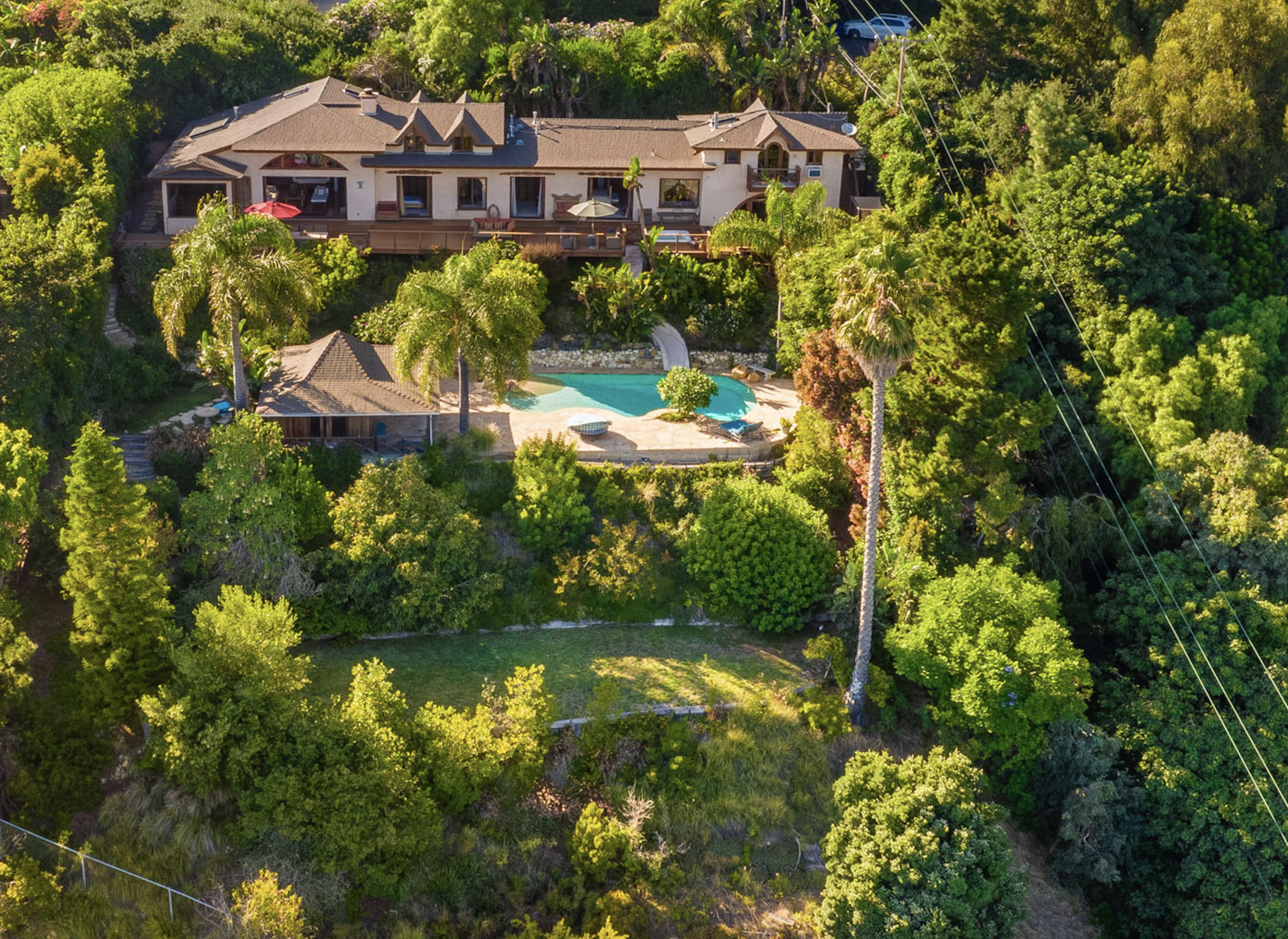 A large house is situated on a hillside surrounded by lush greenery, featuring a pool and a patio area.