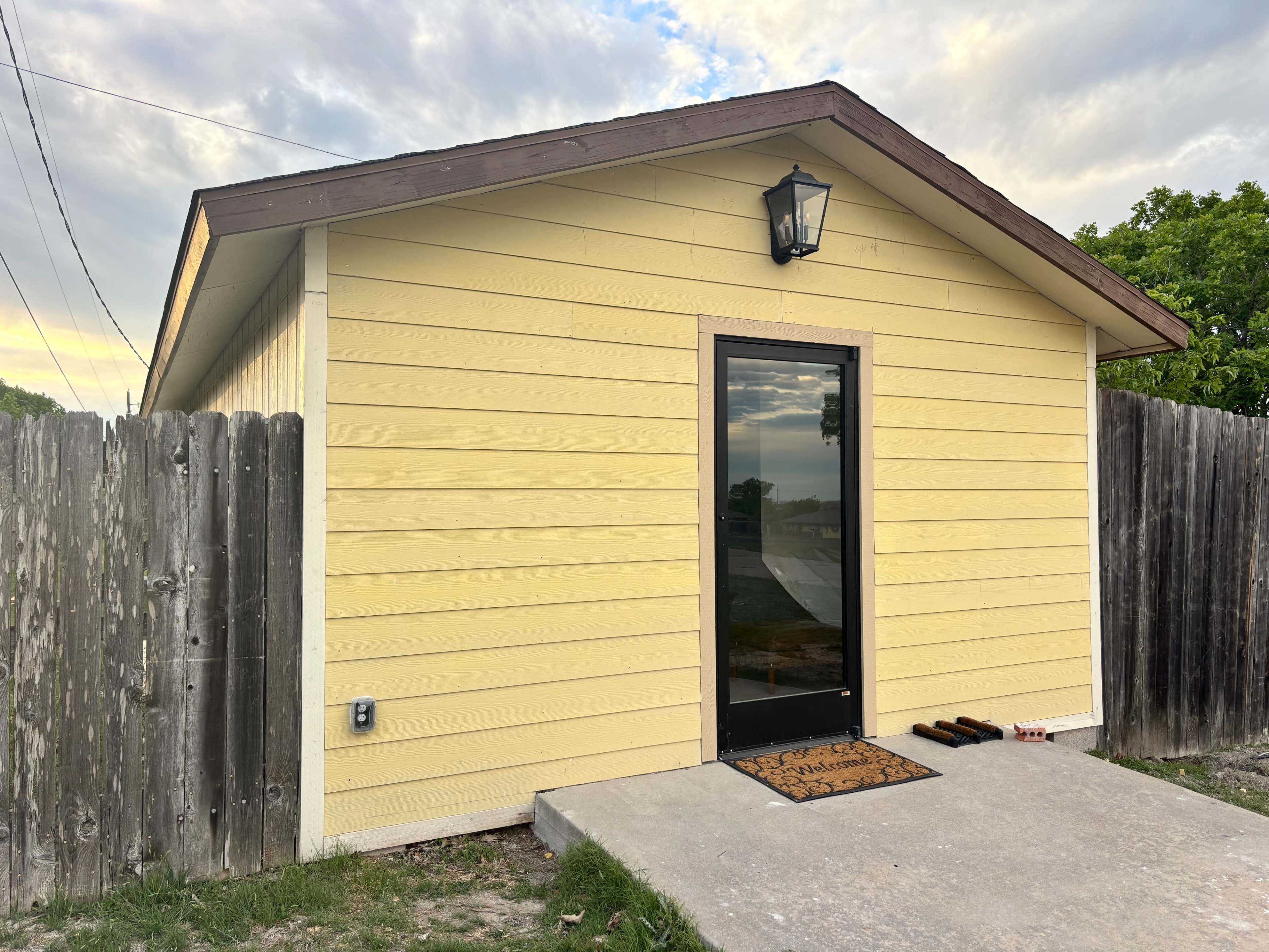 The image shows a yellow house with a black door and a lantern above it, surrounded by a wooden fence and a concrete pathway.