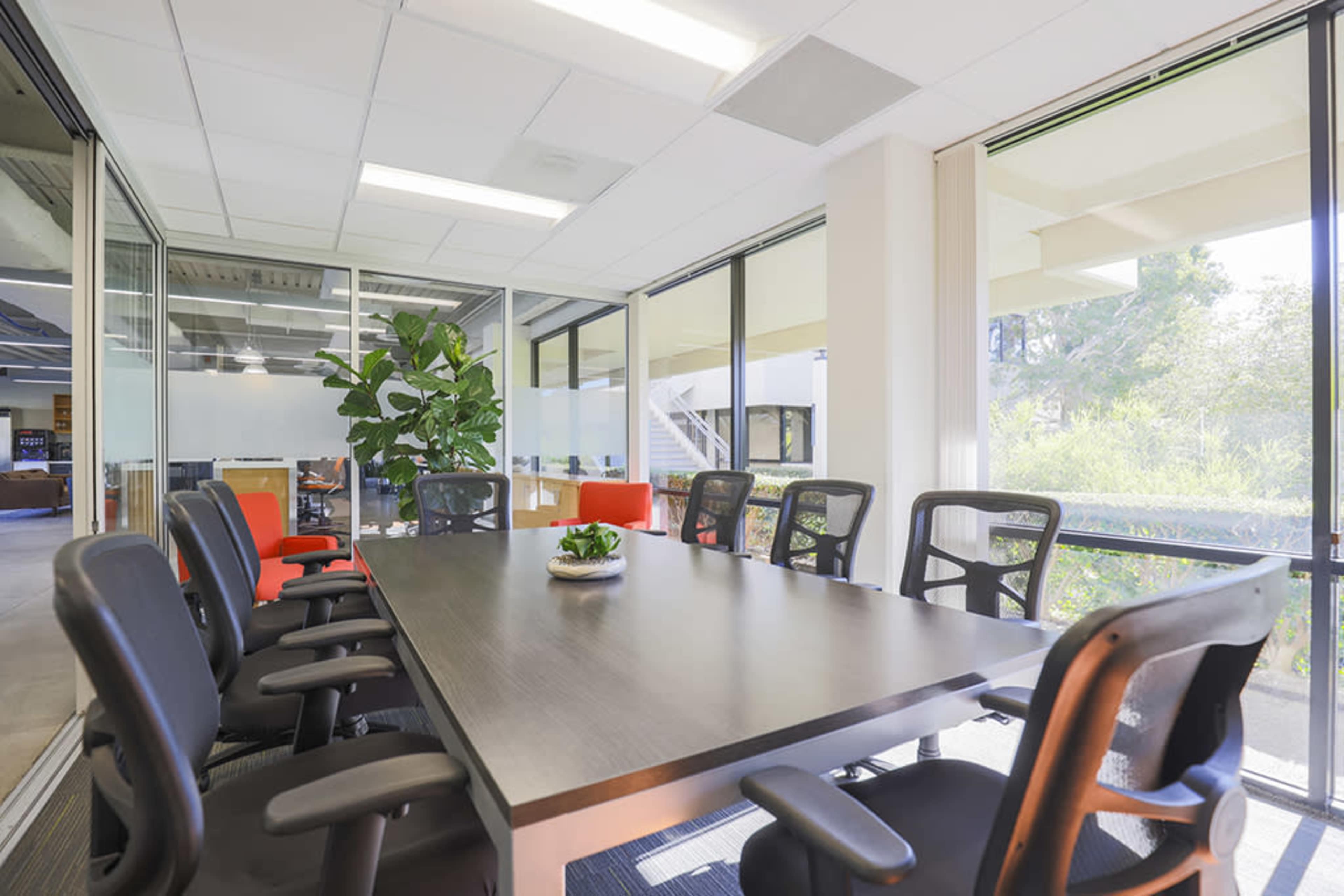 A modern conference room features a long table surrounded by black chairs, with large windows allowing natural light and a view of greenery outside.