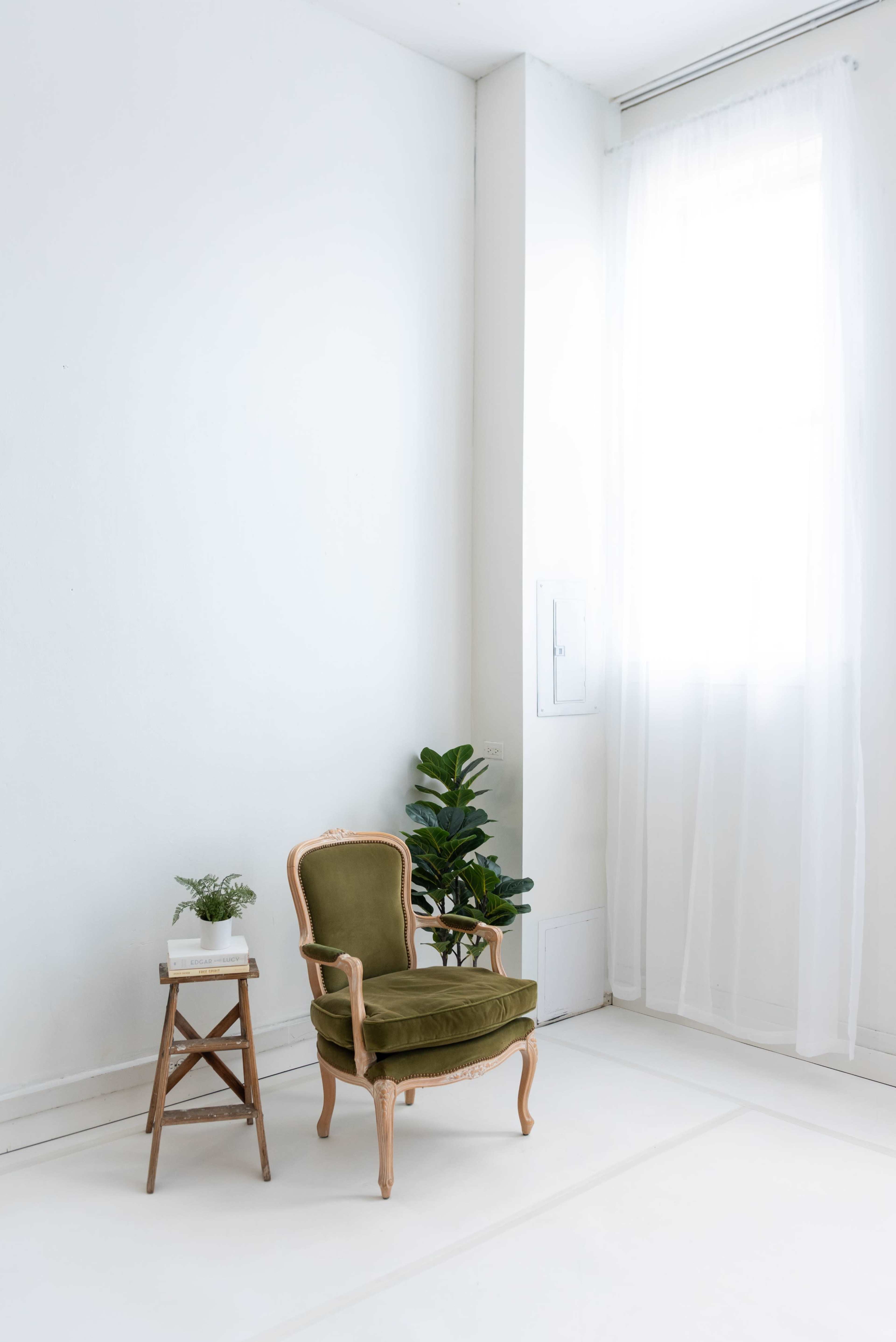 The image shows a simple interior featuring an olive green armchair next to a small wooden table with a potted plant, illuminated by natural light from a window.