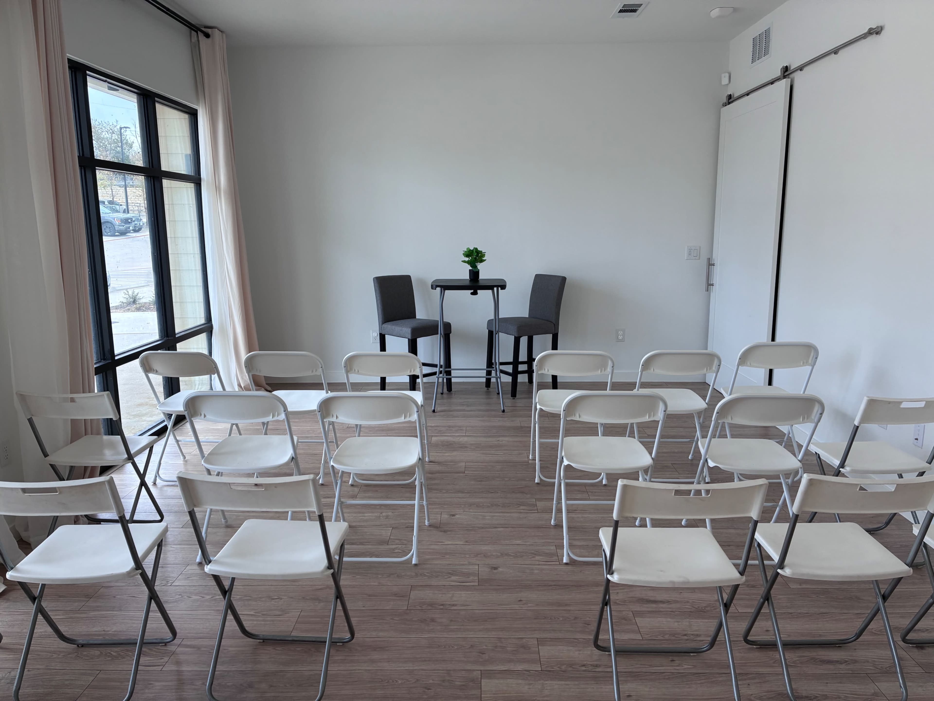 A modern, minimalist room is set up with several rows of folding chairs facing two tall stools and a small table, with a single plant in the center.