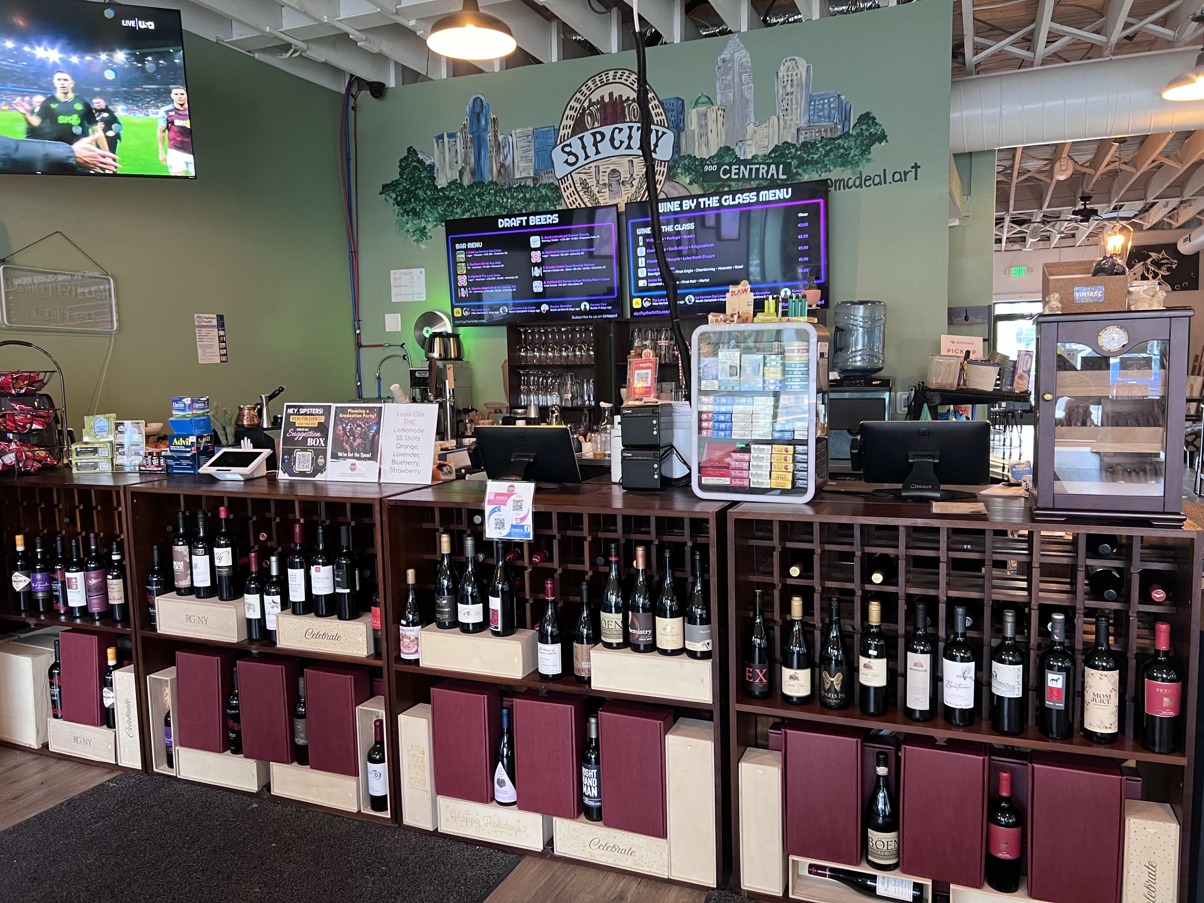 The image shows a bar counter lined with various bottles of wine, accompanied by a menu board displaying drink options and event details in a brightly decorated space.