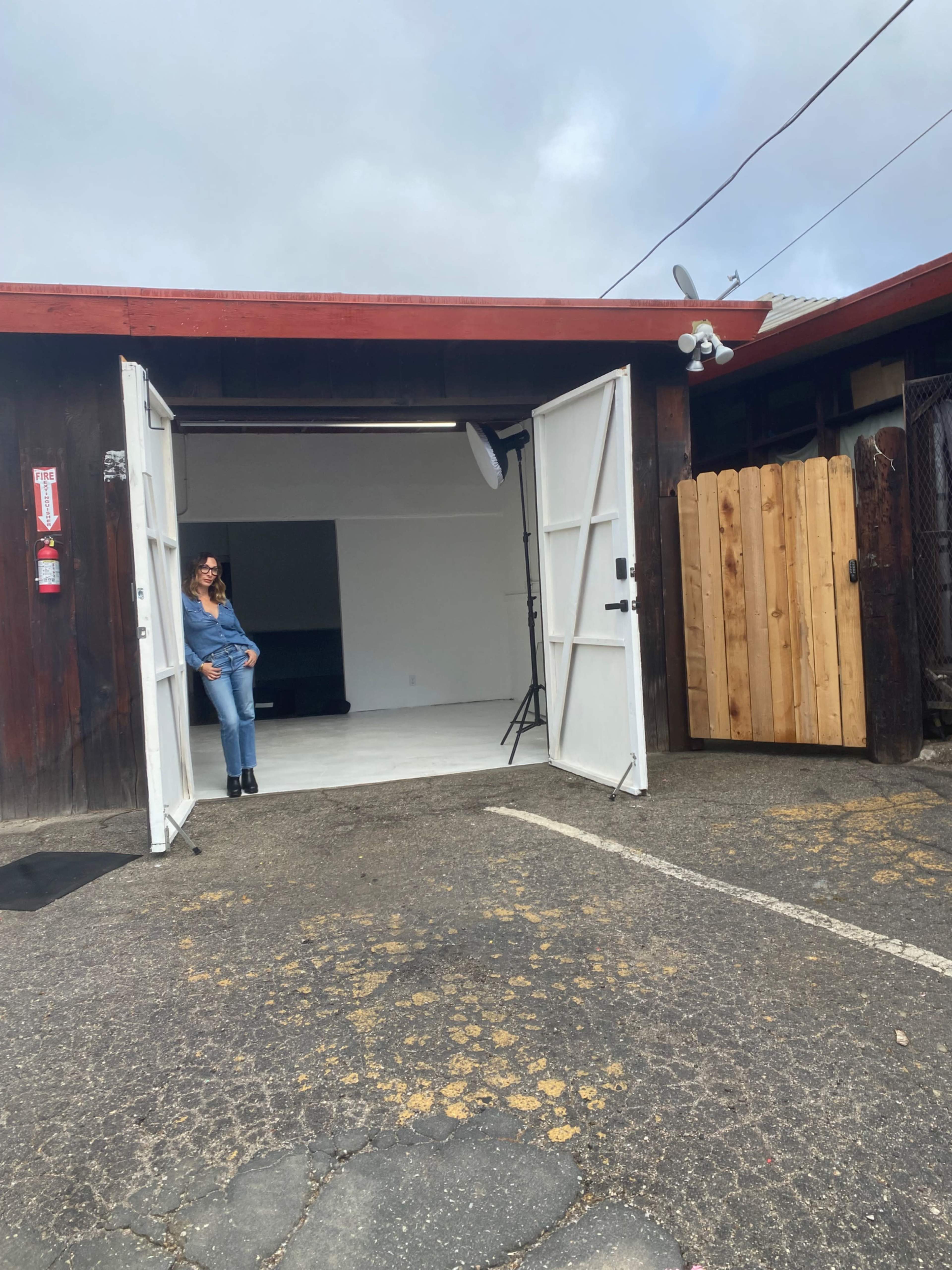 A woman stands in the doorway of a studio with open double doors, revealing a white backdrop and lighting equipment inside.