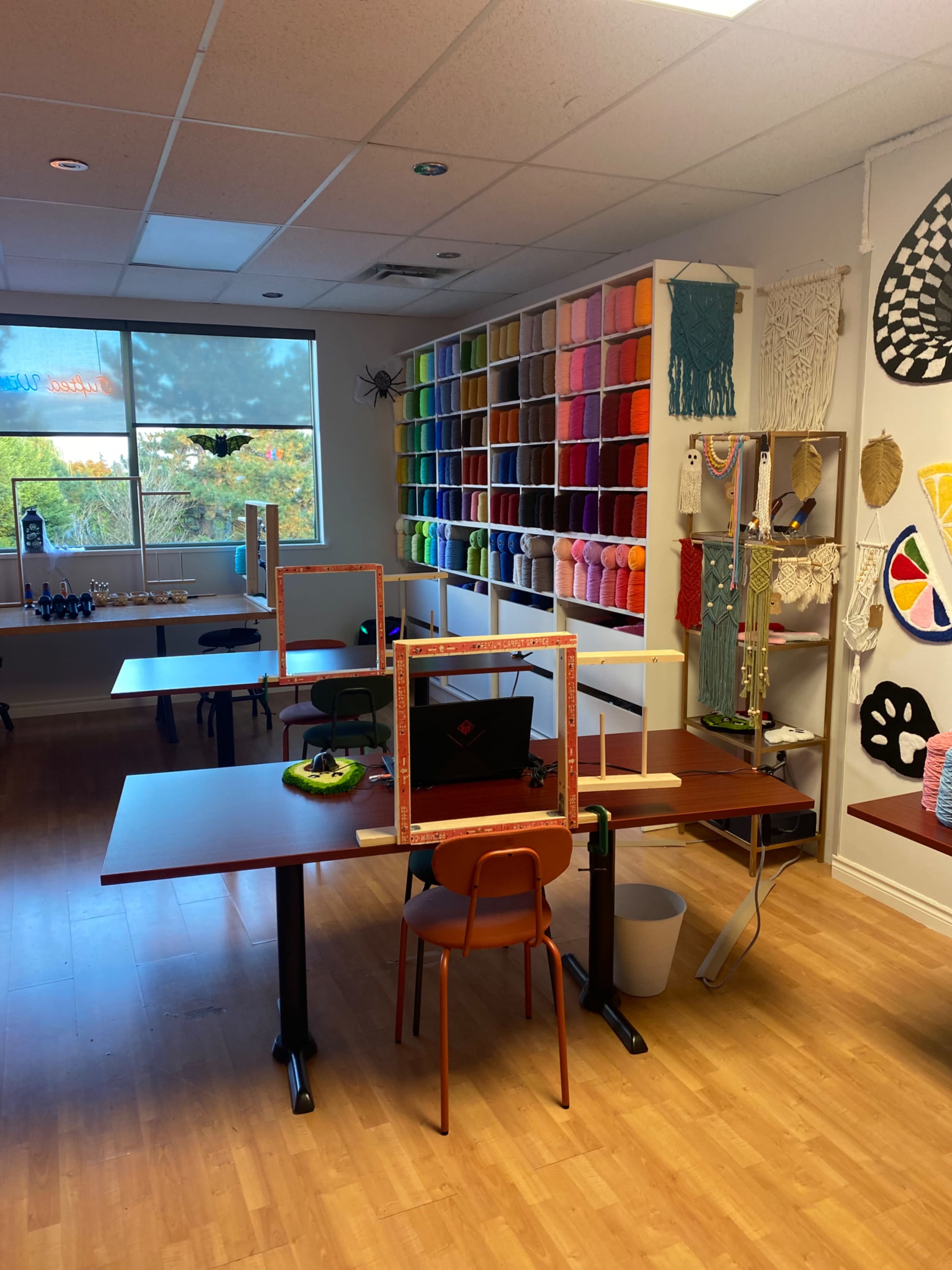The image shows a brightly lit craft room with tables set up for projects, colorful yarn displayed on shelves, and various art supplies decorating the walls.