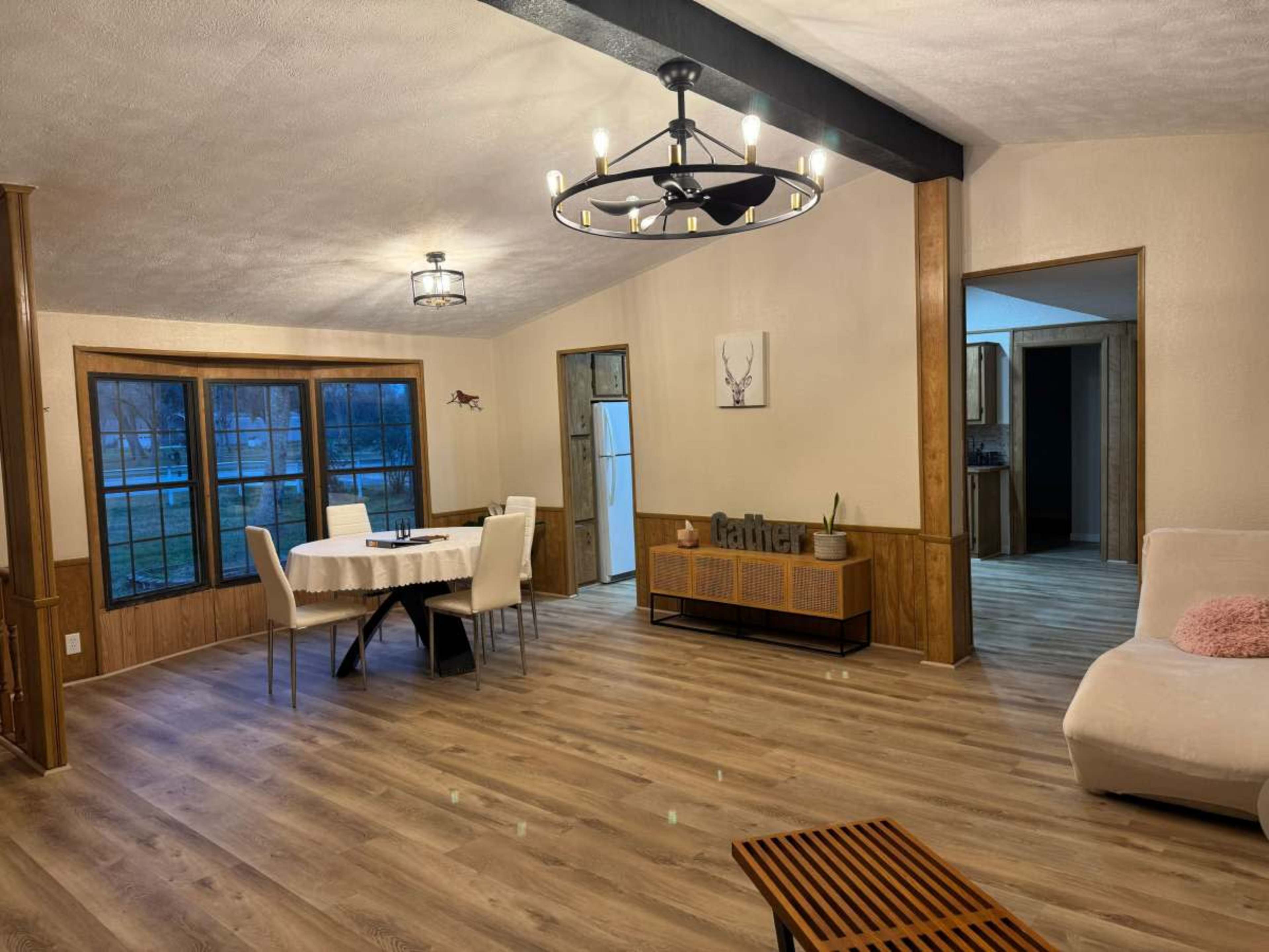 The image shows a modern, open living space with a dining table and chairs, natural light coming through large windows, and a cozy seating area.