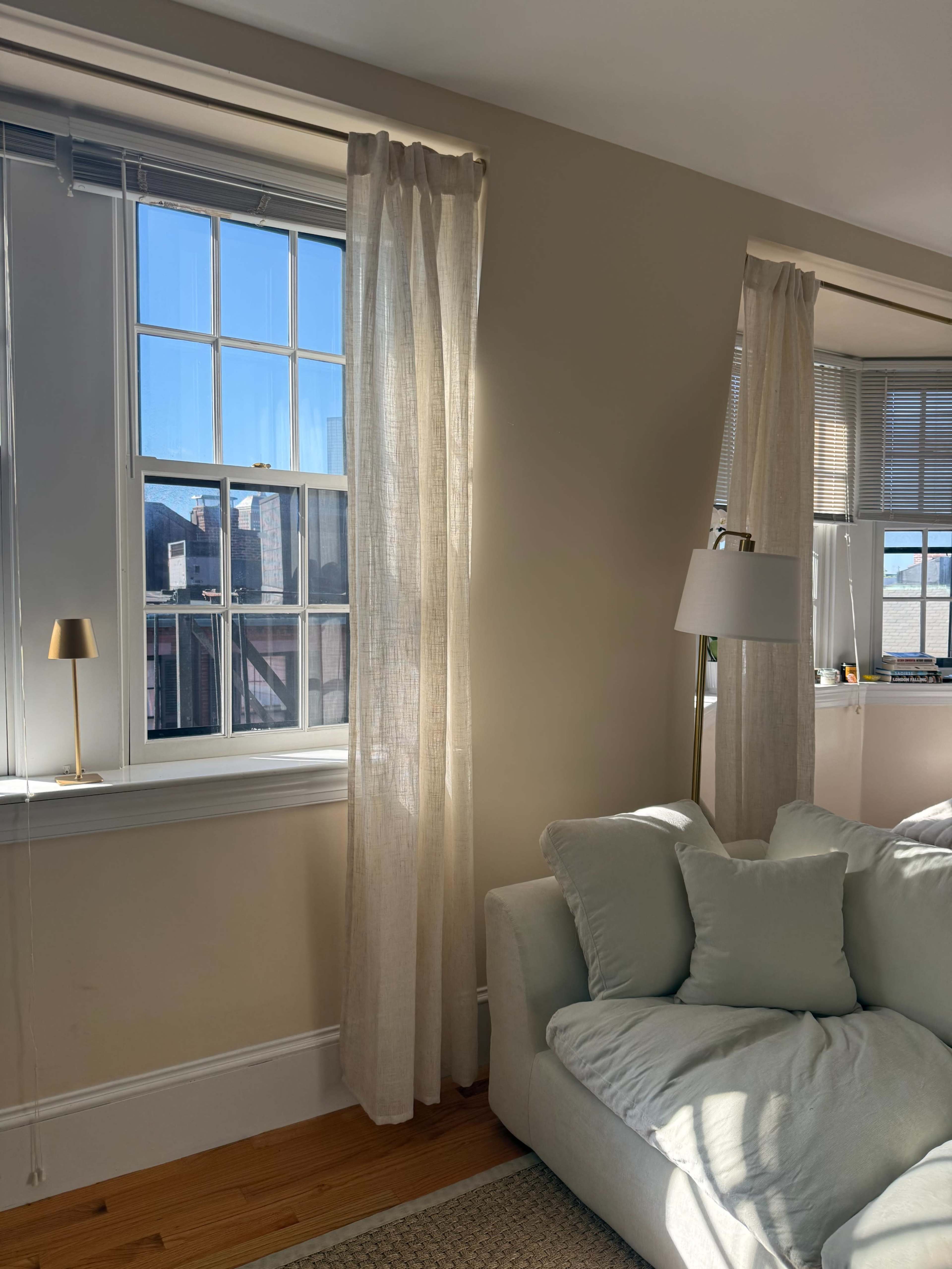 The image shows a sunlit living room with a soft-textured sofa, light curtains, and a window revealing blue skies.