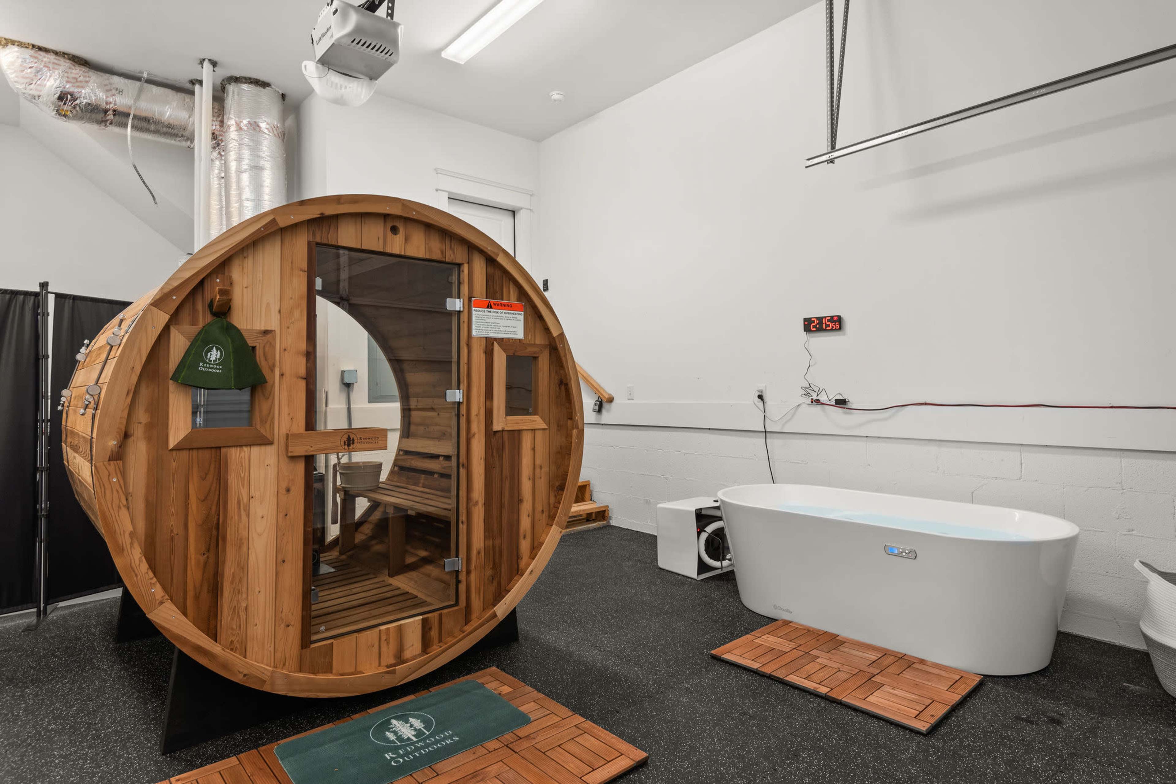 The image shows a wooden sauna barrel next to a modern freestanding bathtub in a room with black flooring and exposed ductwork.