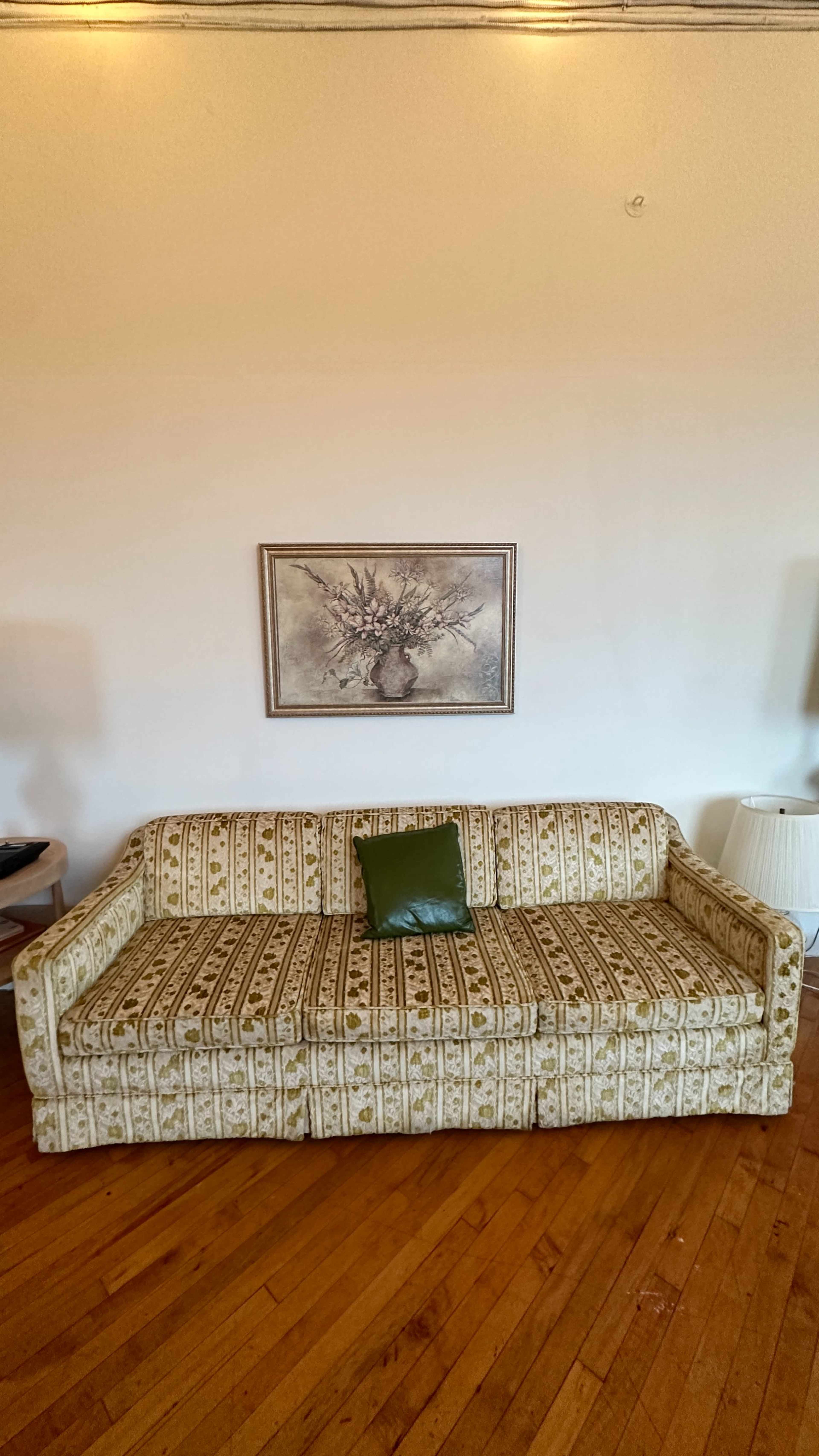A patterned sofa with a green cushion is placed against a white wall, beneath a framed artwork depicting a tree.