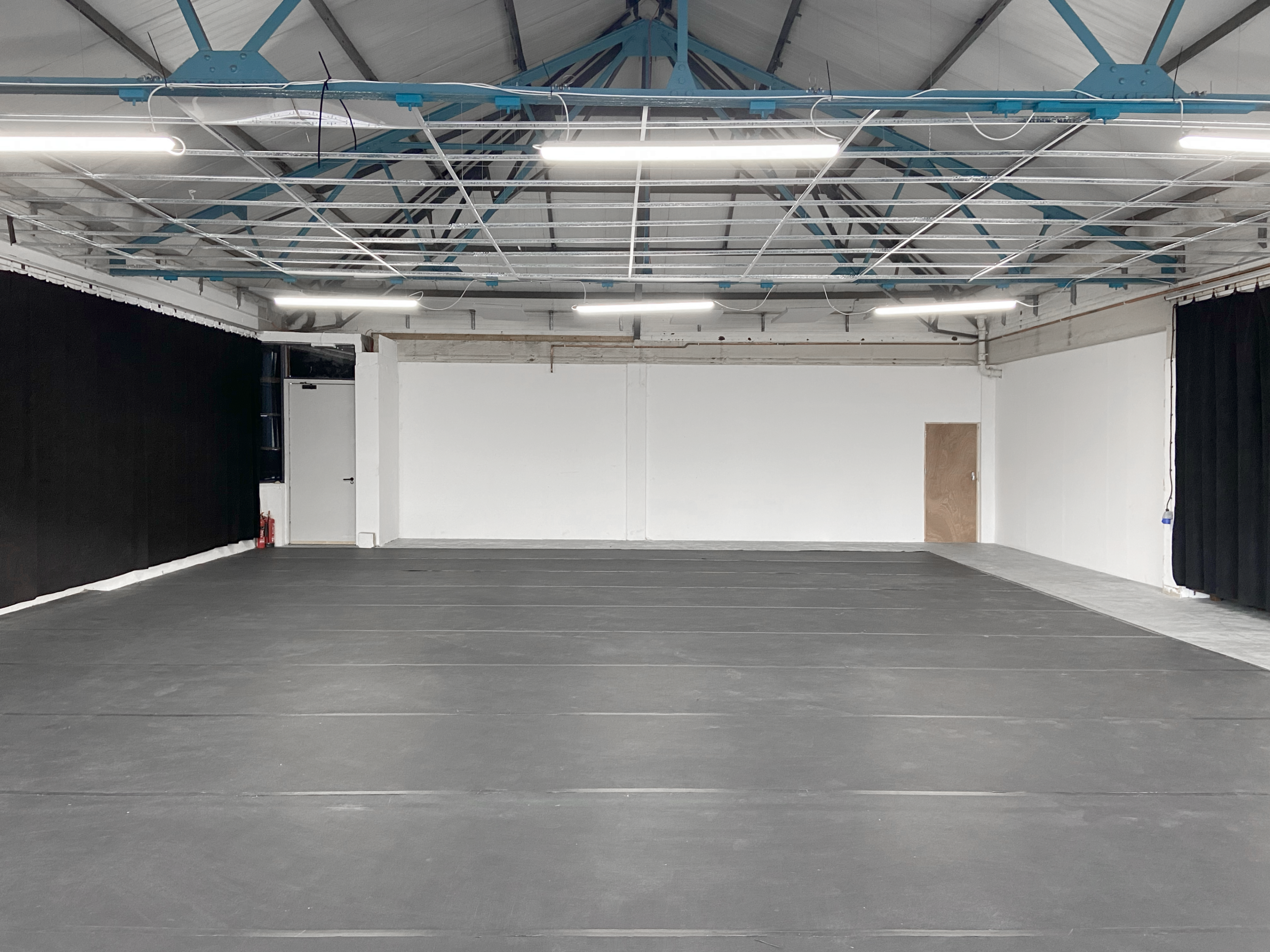 A spacious, empty interior with a black floor, white walls, and a high, exposed ceiling with blue beams.