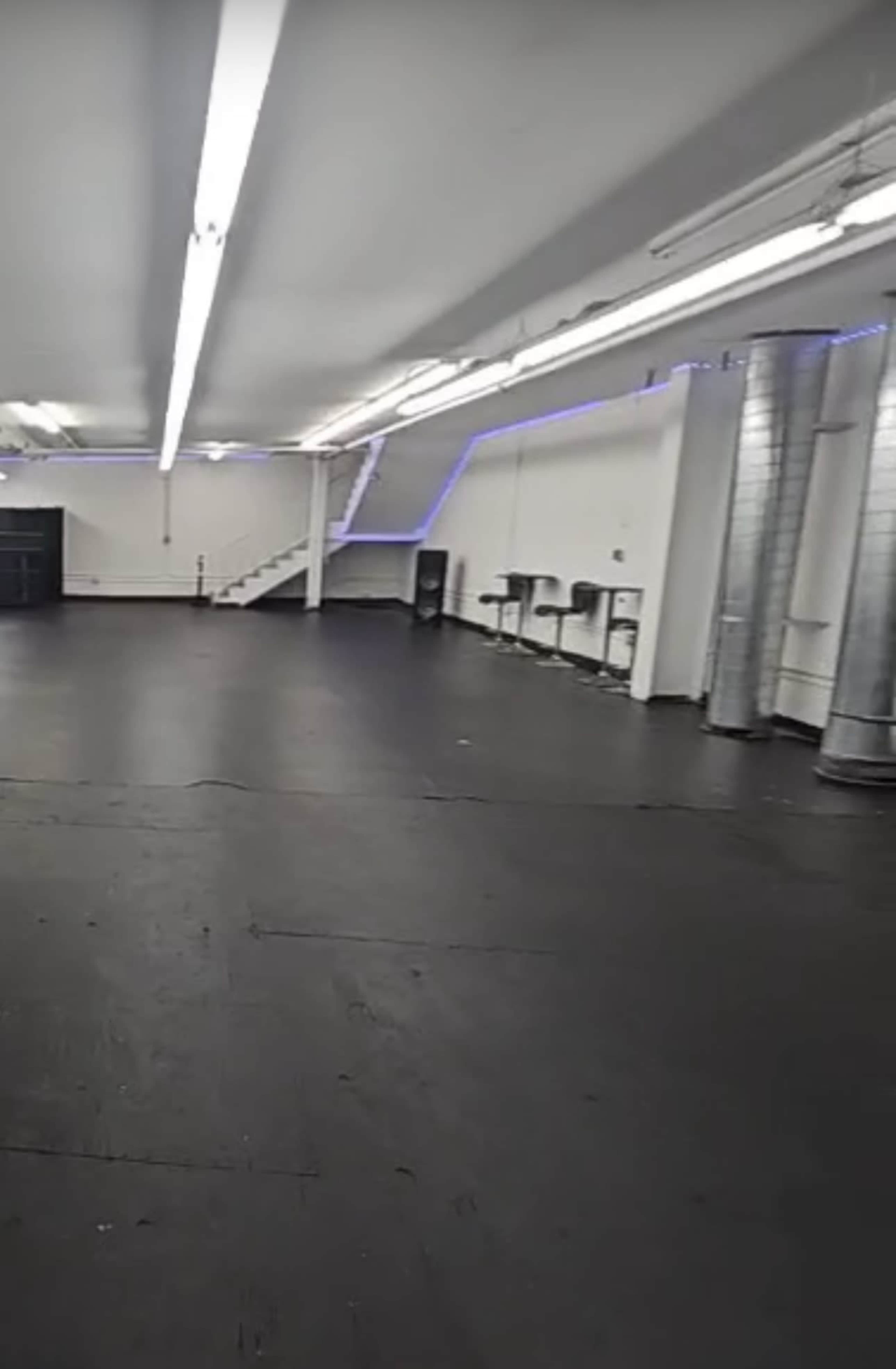 The image shows an empty, spacious room with black flooring, bright overhead lights, and a staircase leading to a higher level in the background.