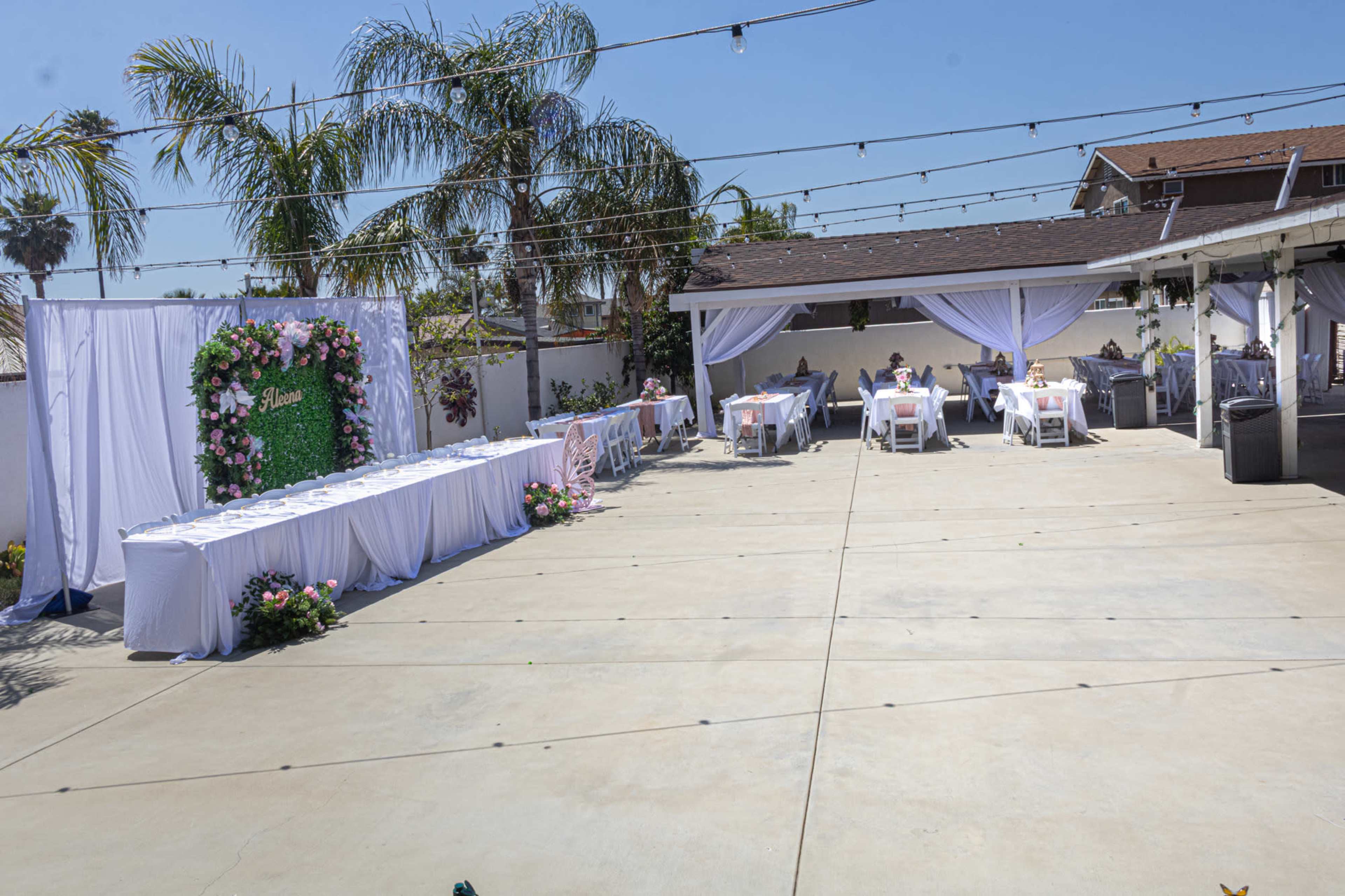 The image shows a spacious outdoor venue set up for an event, featuring decorated tables and a floral backdrop, with palm trees and string lights overhead.