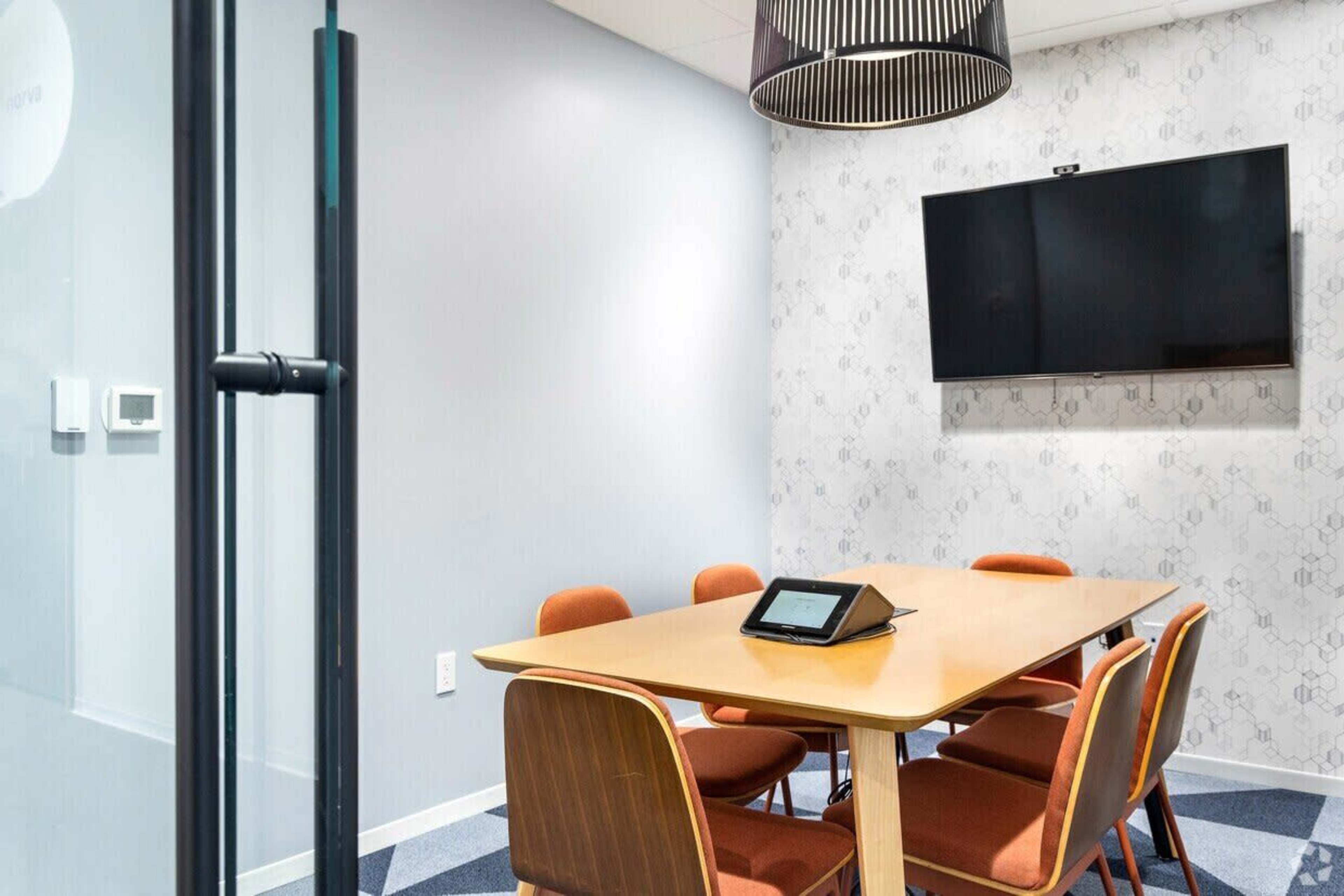 A meeting room features a wooden table surrounded by six orange chairs, a wall-mounted television, and a touchscreen device on the table.