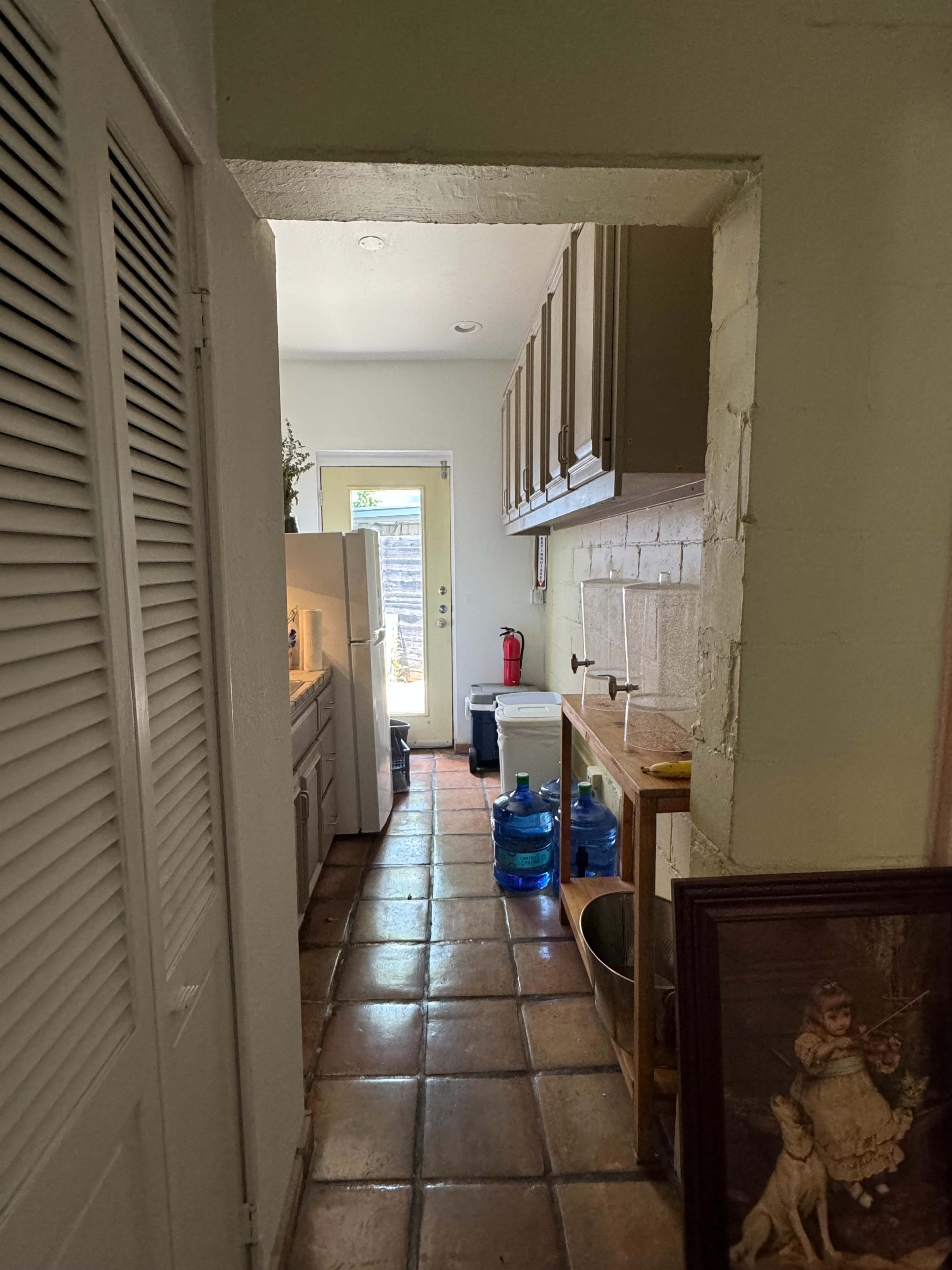 A narrow hallway connects a kitchen to an outdoor area, featuring cabinets on one side, appliances, and several large water bottles.