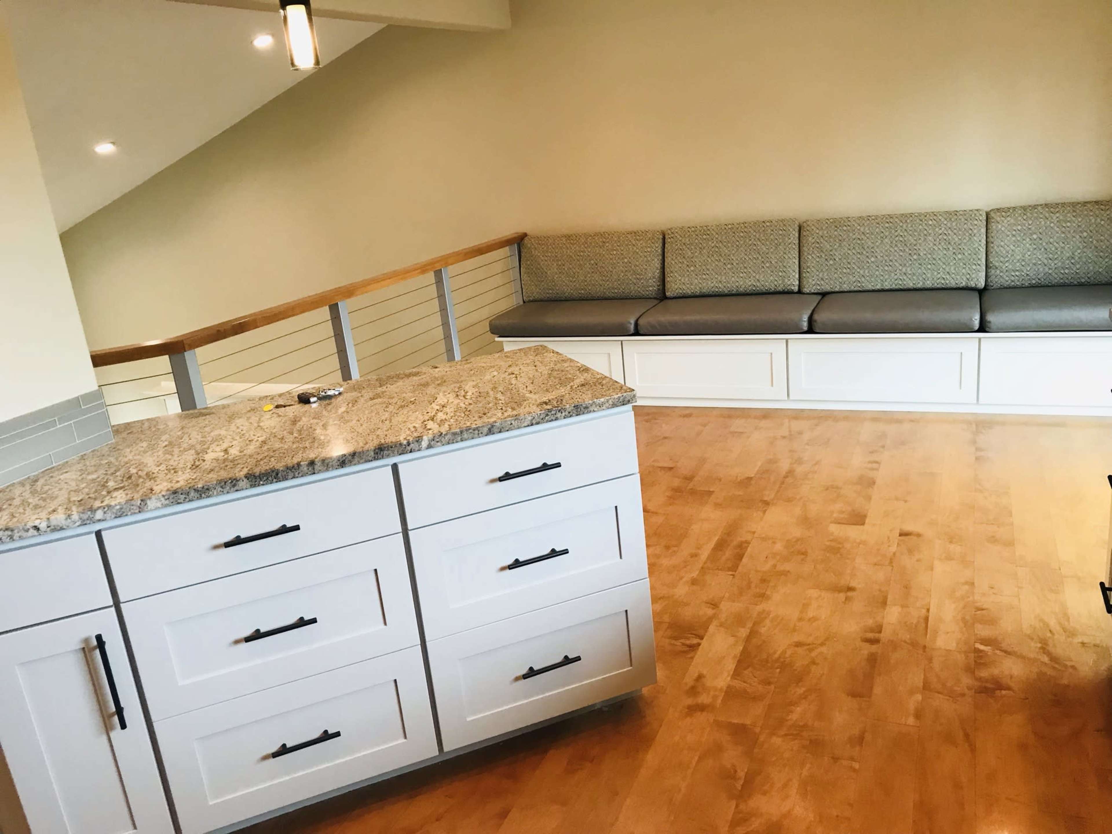 A modern interior featuring a kitchen space with a granite countertop and white cabinetry, alongside a built-in bench with gray cushions against a wall.