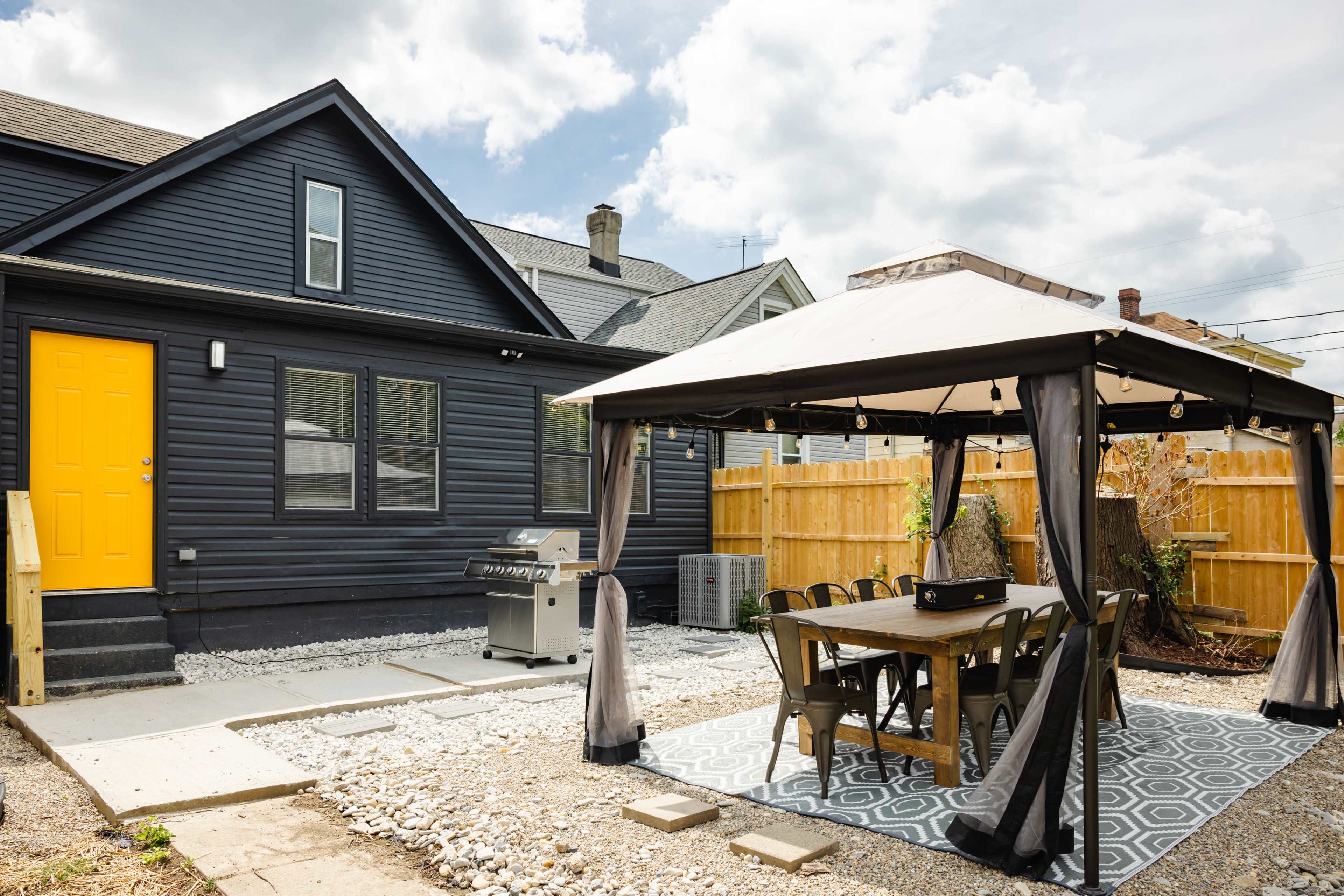 A backyard features a tented dining area next to a grill and a house with a bright yellow door.
