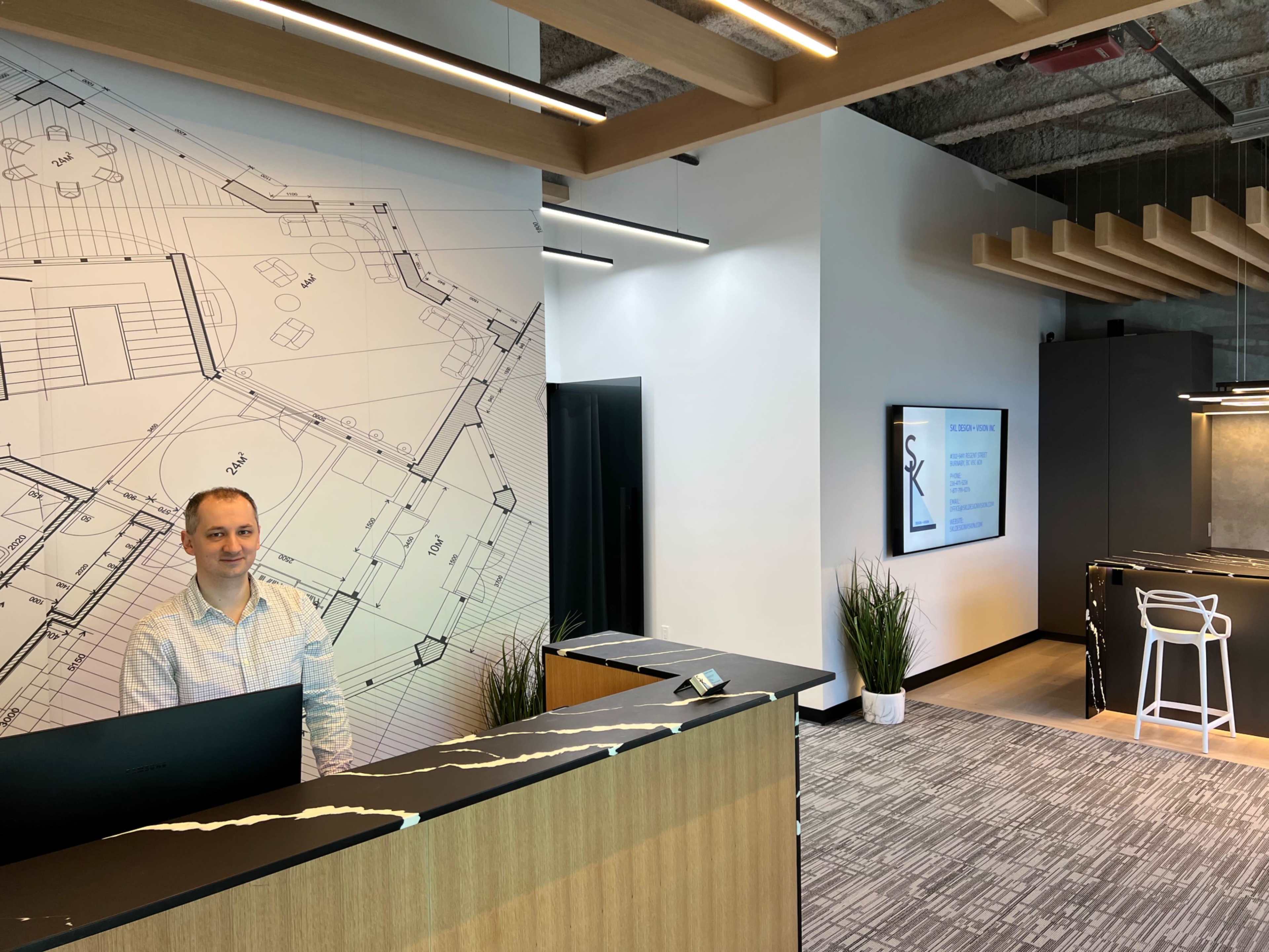 A reception area features a front desk with a blueprint mural on the wall and a screen displaying information in the background.