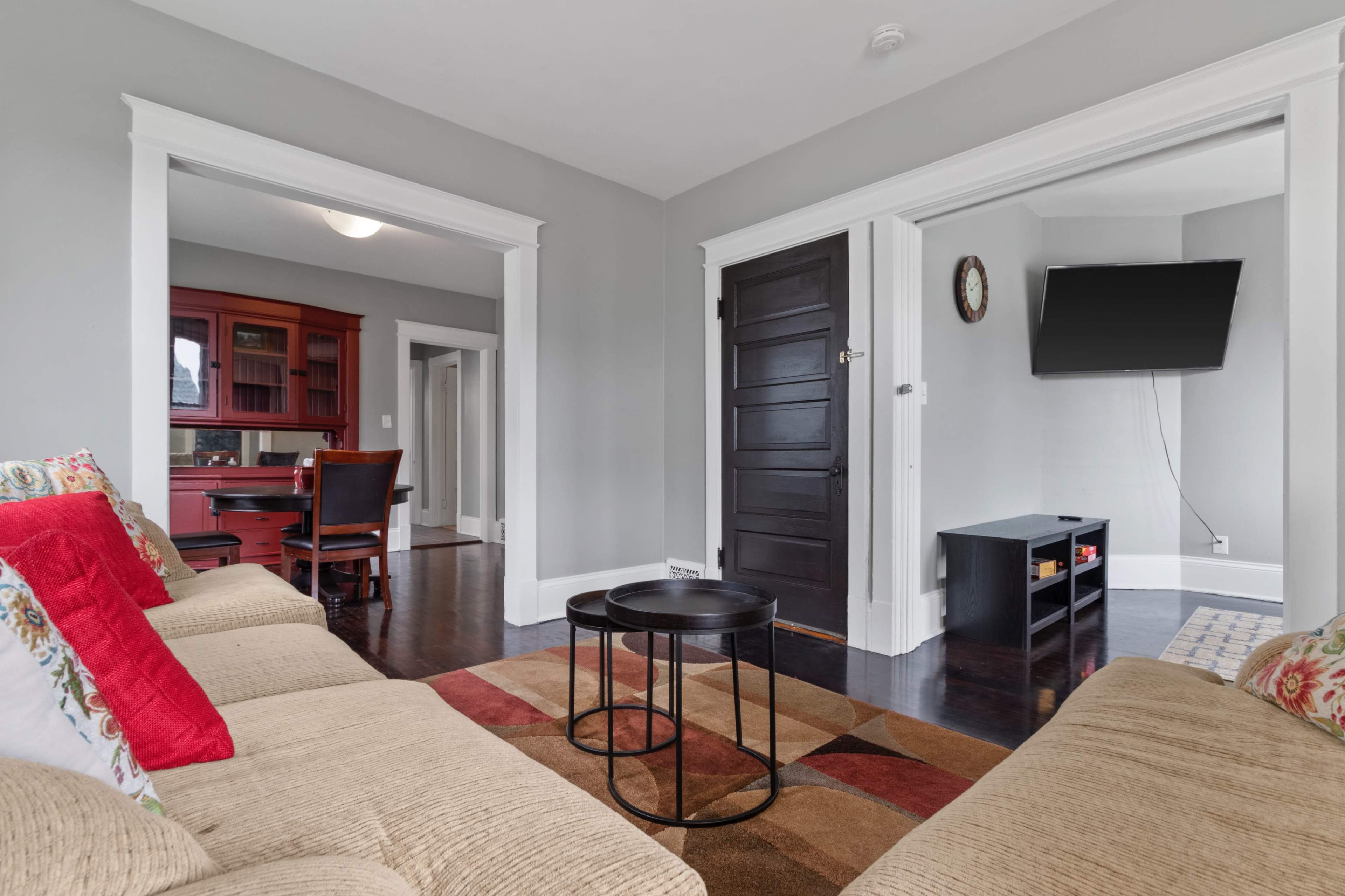 A living area with a tan couch, a small black coffee table, and a wall-mounted TV, leading into a dining area with a red cabinet and wooden table.