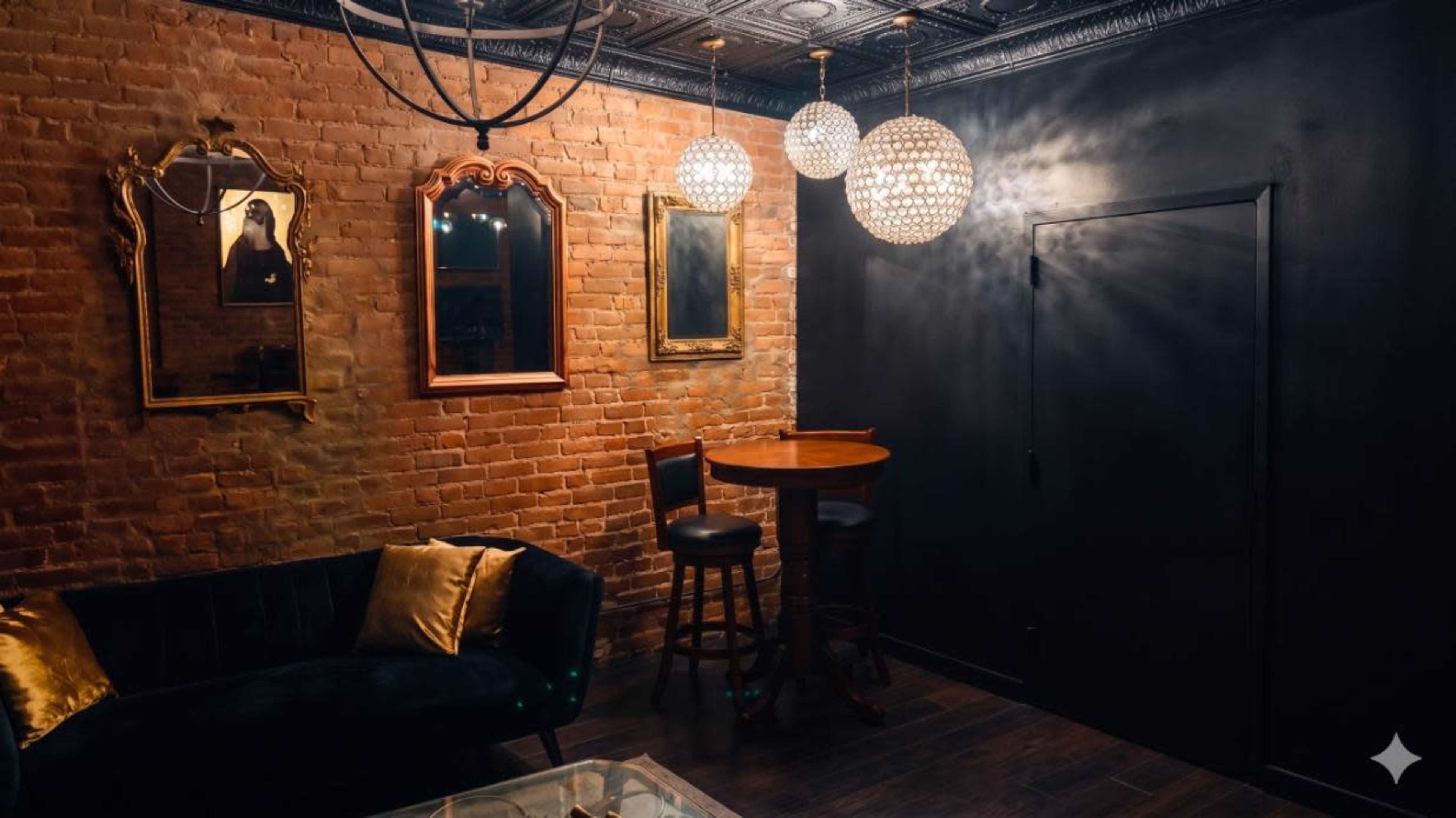 The image shows a dimly lit room with exposed brick walls, a circular wooden table with two stools, and three hanging crystal light fixtures, along with framed mirrors on the wall.