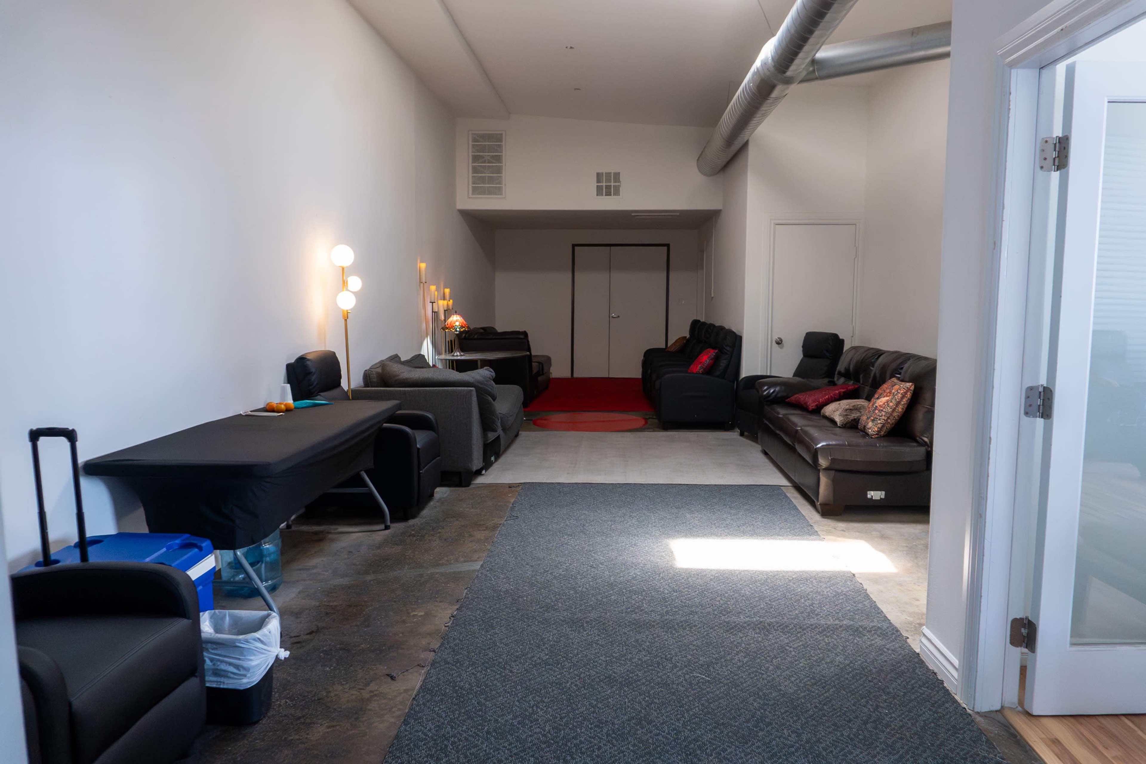 The image shows a long, narrow room featuring a row of black leather sofas on one side, a dark table with some items on it, and a gray carpet running along the floor.