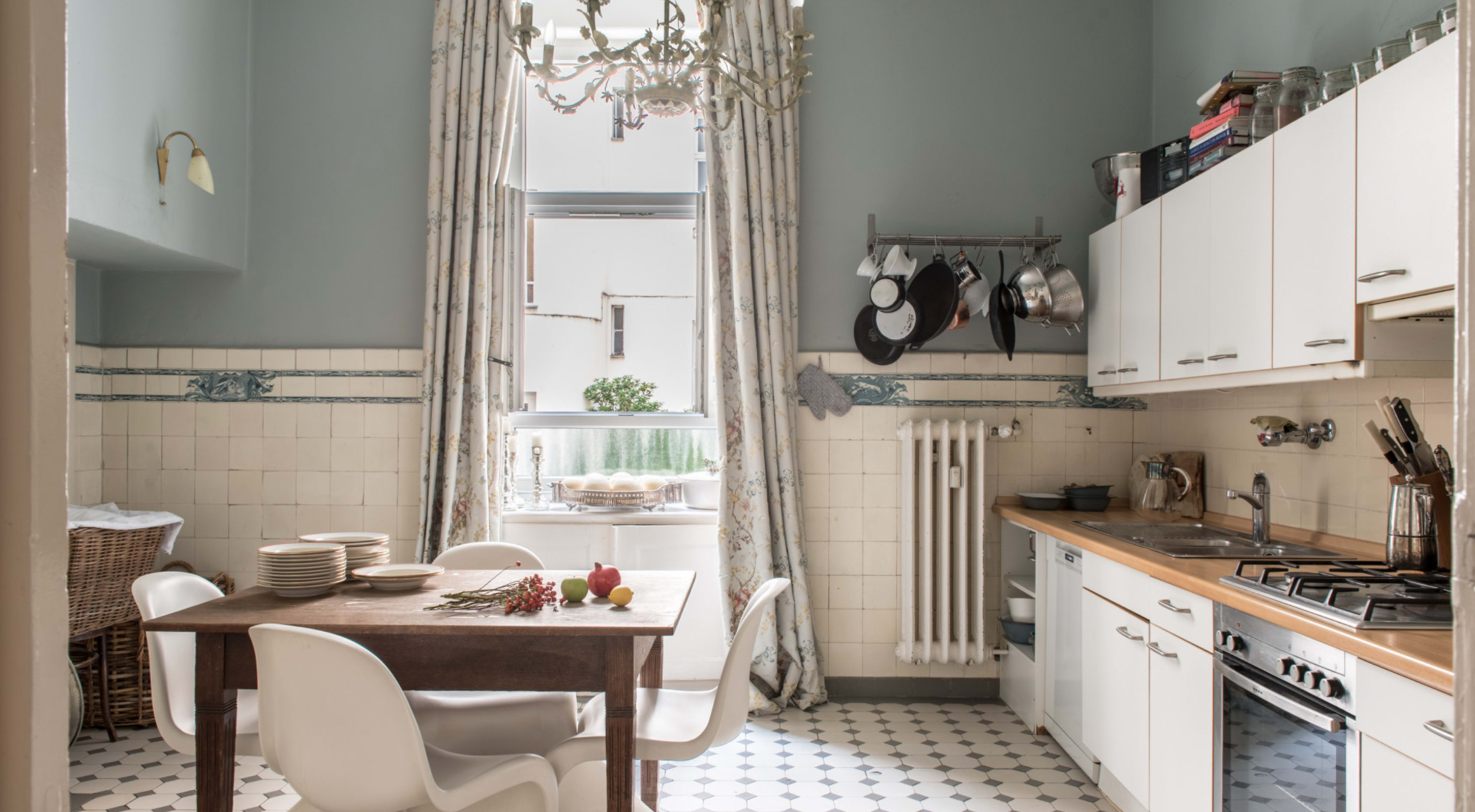 The kitchen features a wooden table with white chairs, a window with patterned curtains, and stainless steel cookware hanging on a rack.