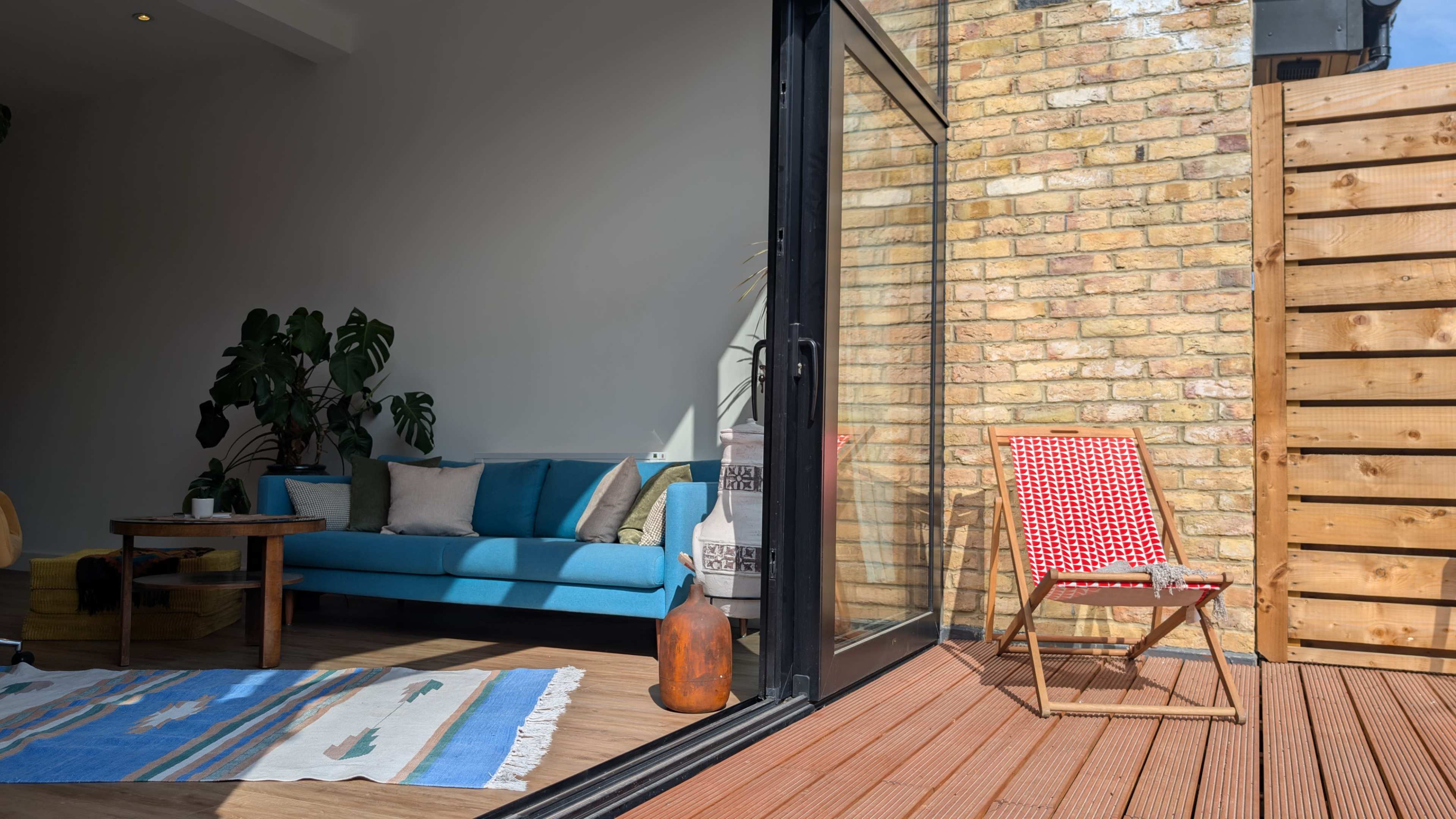 A bright living space features a blue couch, a wooden table, and a sliding glass door leading to an outdoor deck with a wooden screen and a red-and-white patterned chair.