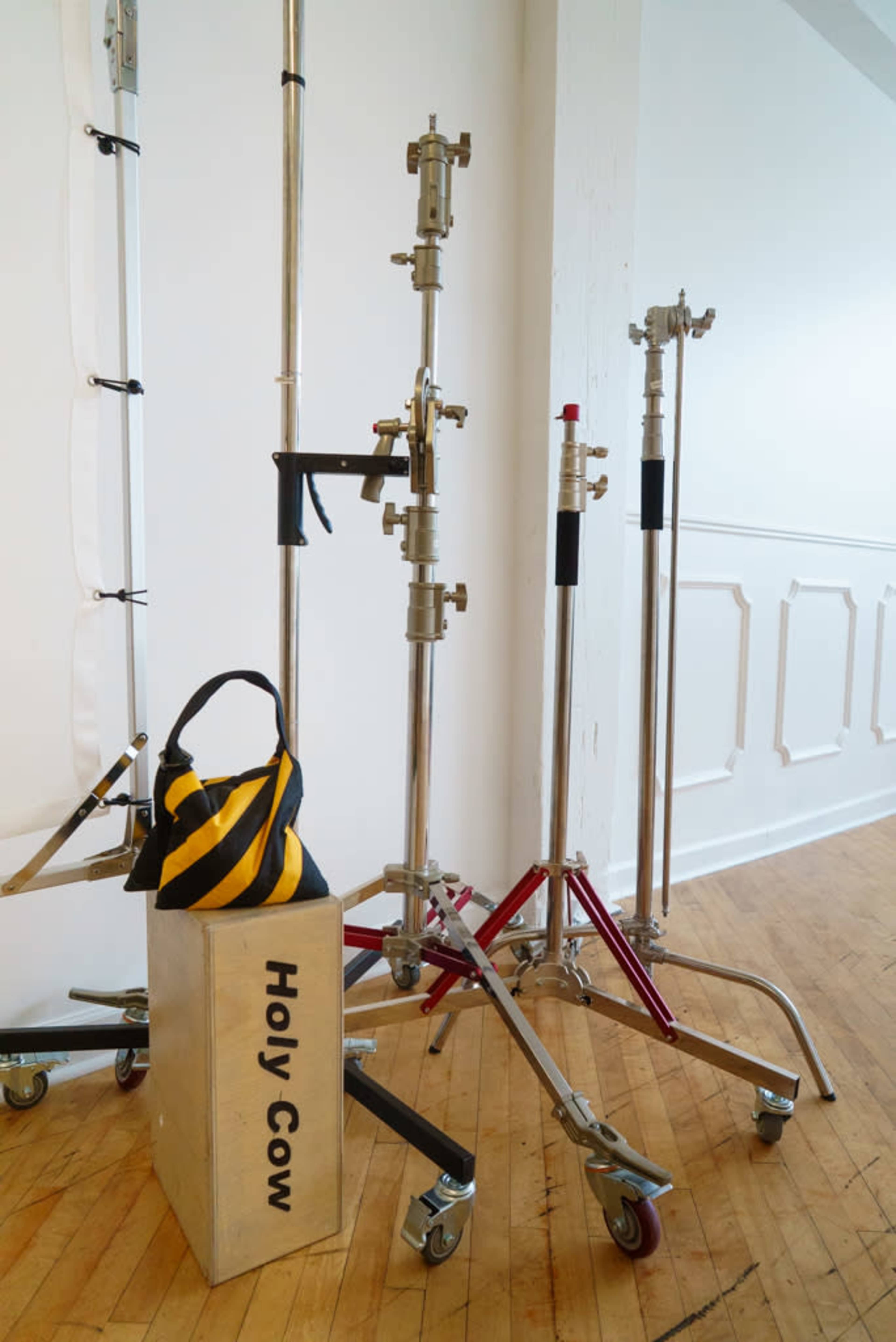The image shows several metal lighting stands and a weighted sandbag labeled "Holy Cow" on a wooden floor in a bright, minimalistic space.