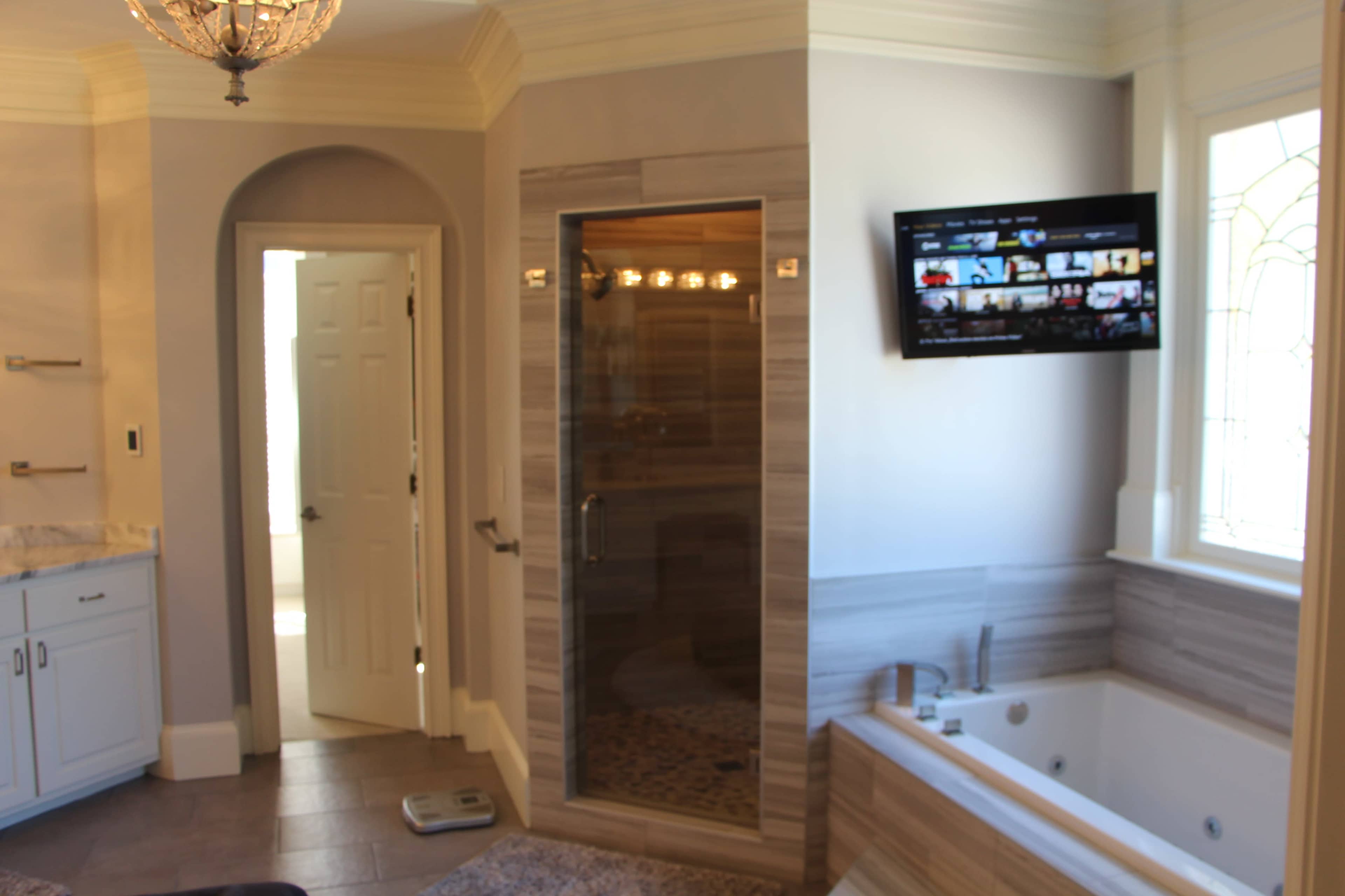 The image shows a spacious bathroom featuring a glass shower, a jetted bathtub, and a flat-screen TV mounted on the wall.