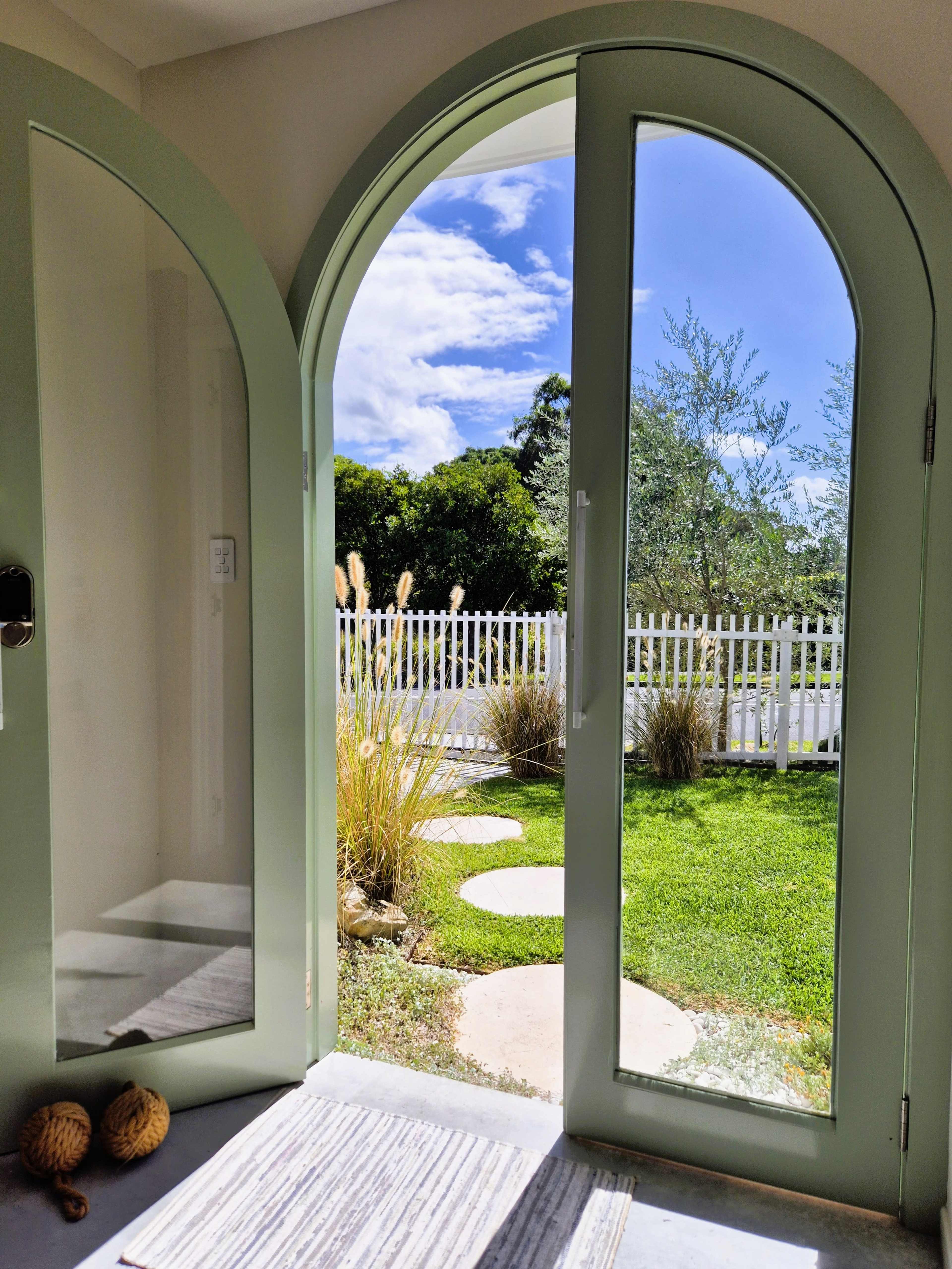 A pair of arched green doors opens to a yard with a white picket fence, manicured grass, and decorative plants under a partly cloudy sky.