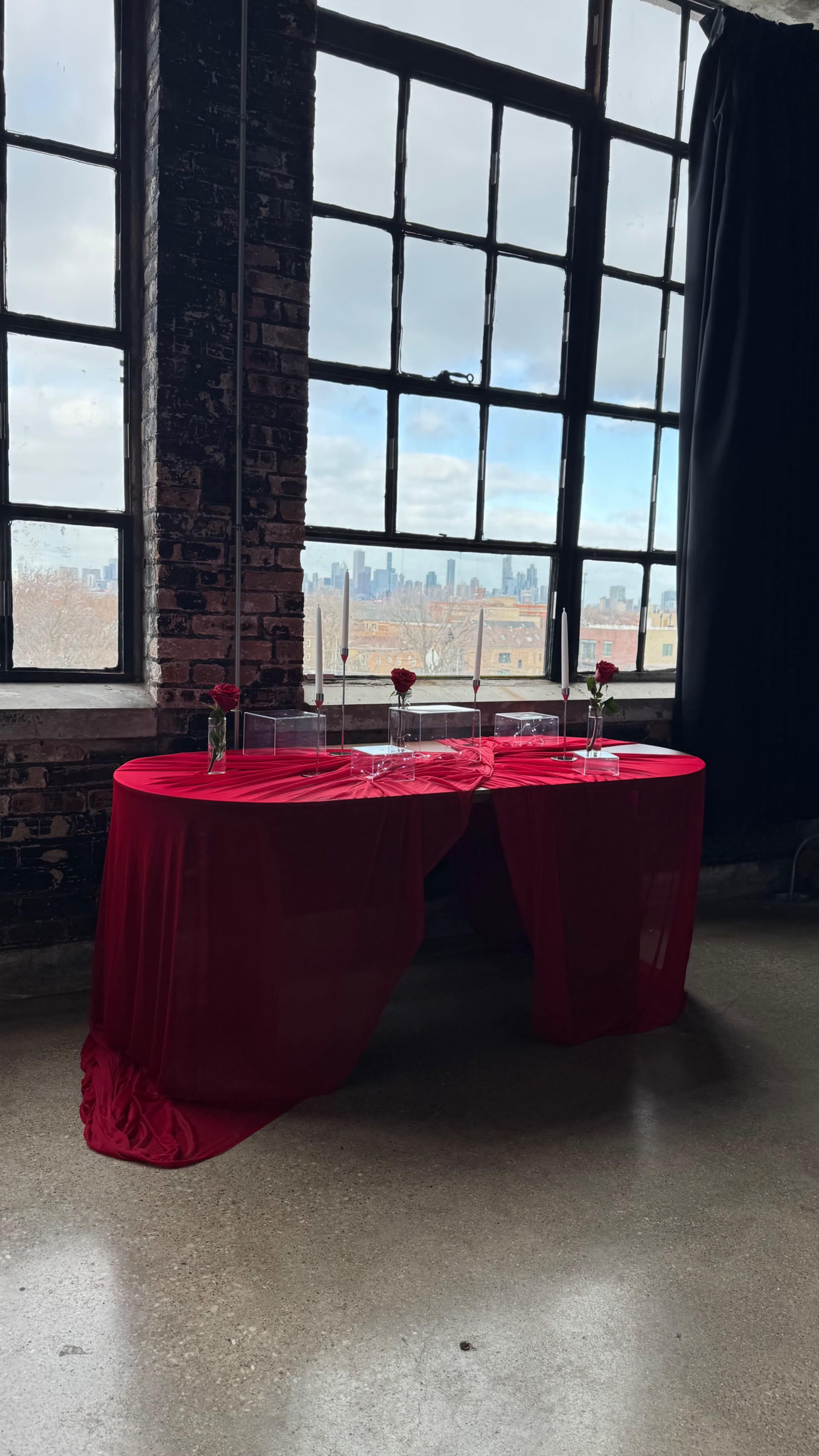 A red-draped table is positioned near large industrial windows that offer a view of the city skyline.