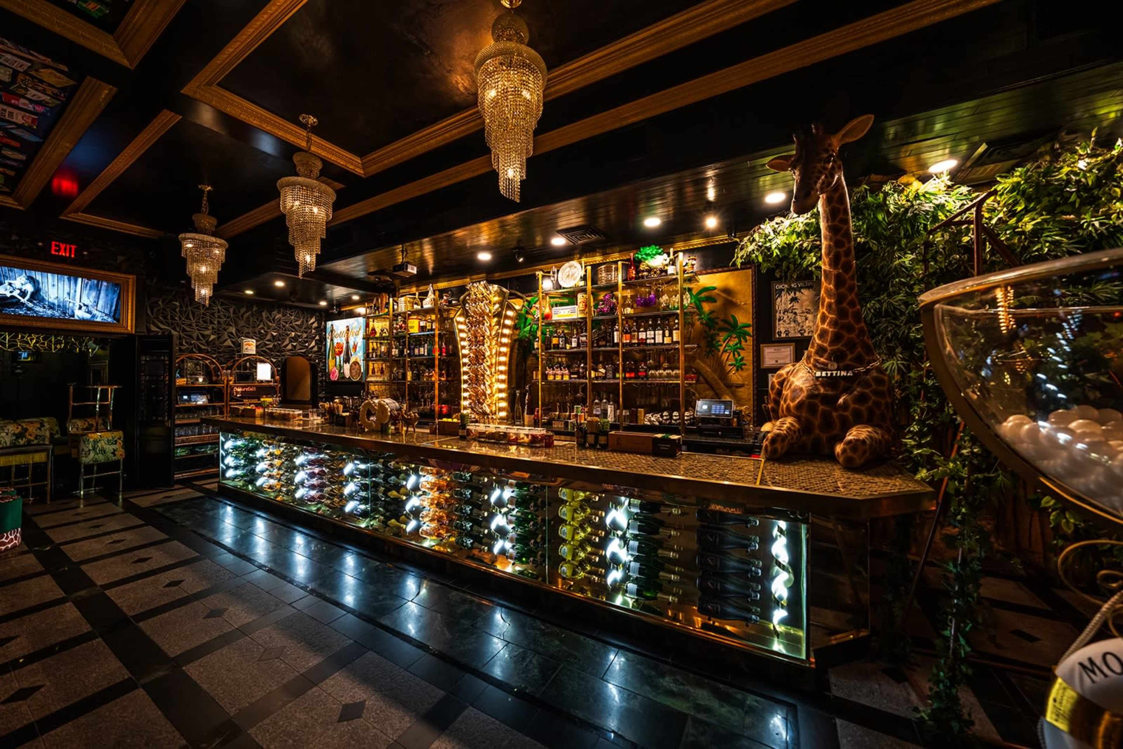 The image shows a vibrant bar with a gold-accented counter, various bottles displayed on shelves, and decorative elements including a giraffe statue and hanging chandeliers.