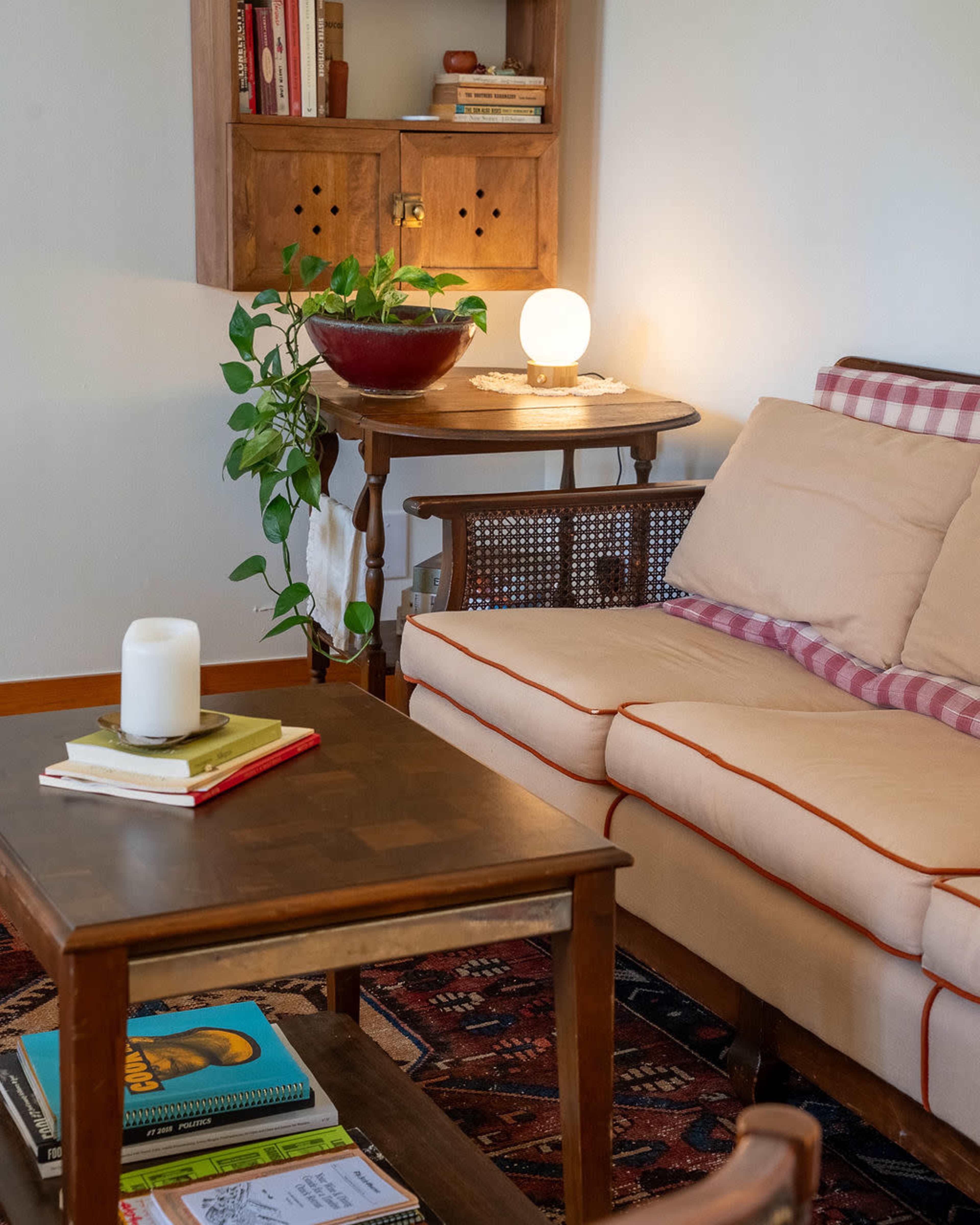 A cozy living room features a beige sofa, a wooden coffee table, a bookshelf with books, a bowl with plants, and a lamp, all arranged on a patterned rug.