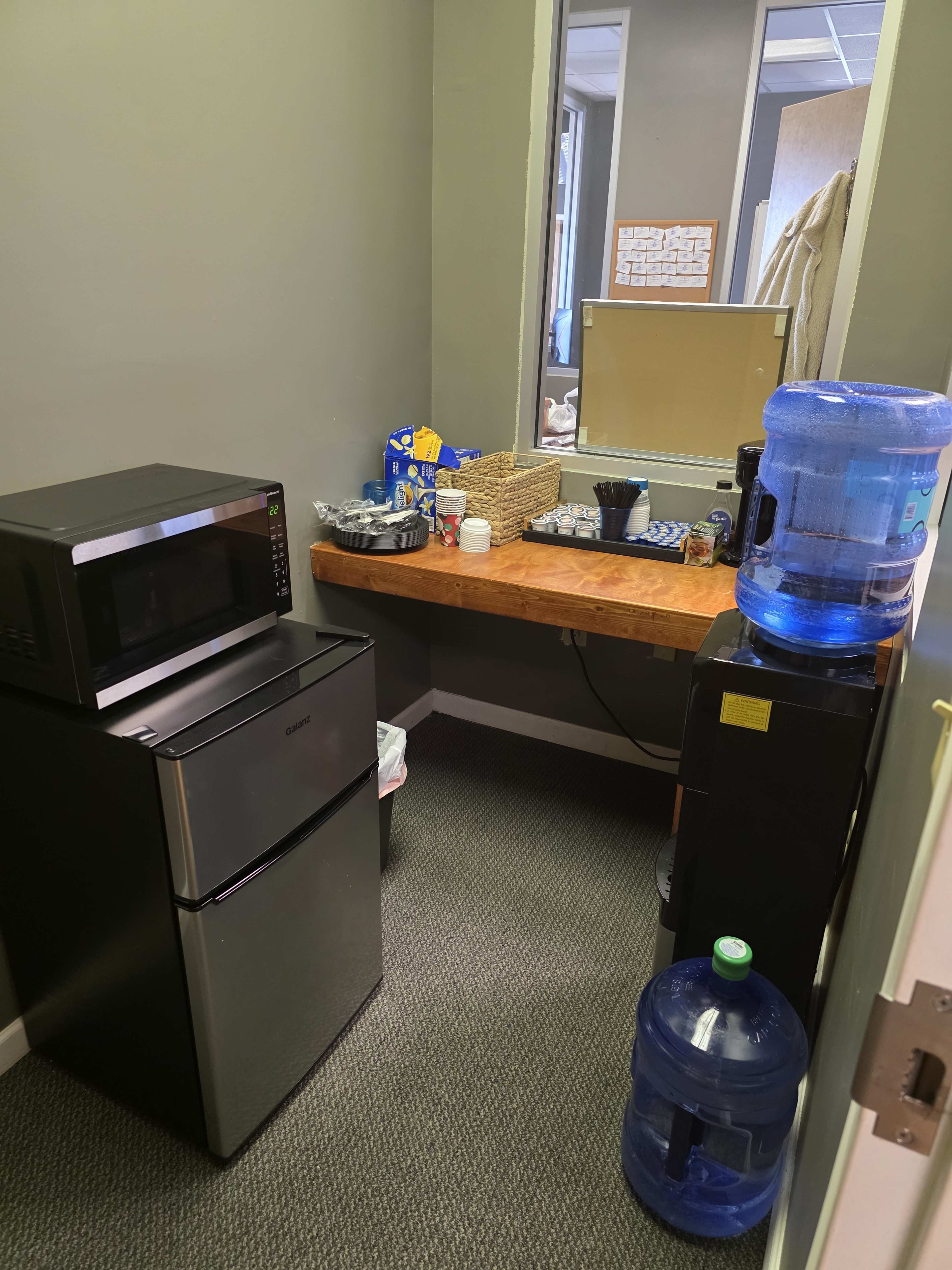 The image shows a small kitchenette with a black microwave, a mini refrigerator, a wooden table with various kitchen items, and a large water cooler.