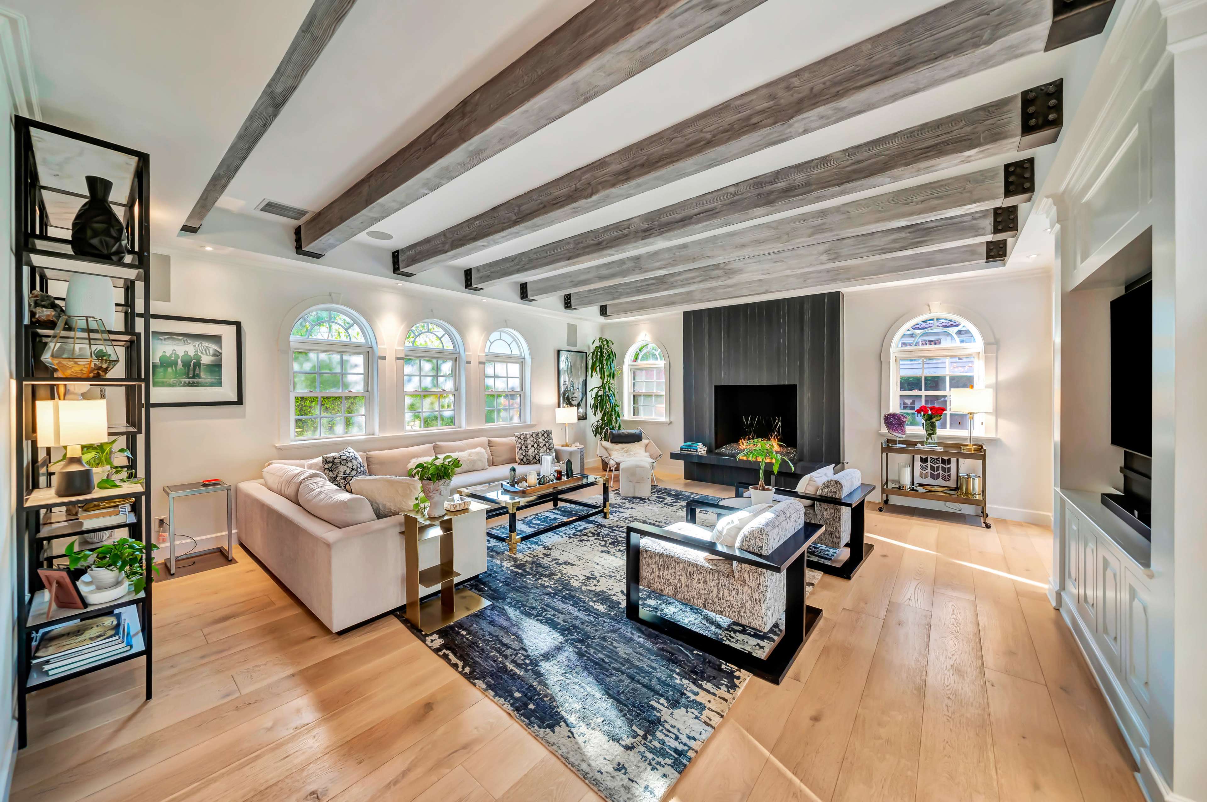 The image shows a modern living room with wooden beams on the ceiling, a large television on the wall, and a seating area with a sofa and chairs arranged around a coffee table on a colorful rug.