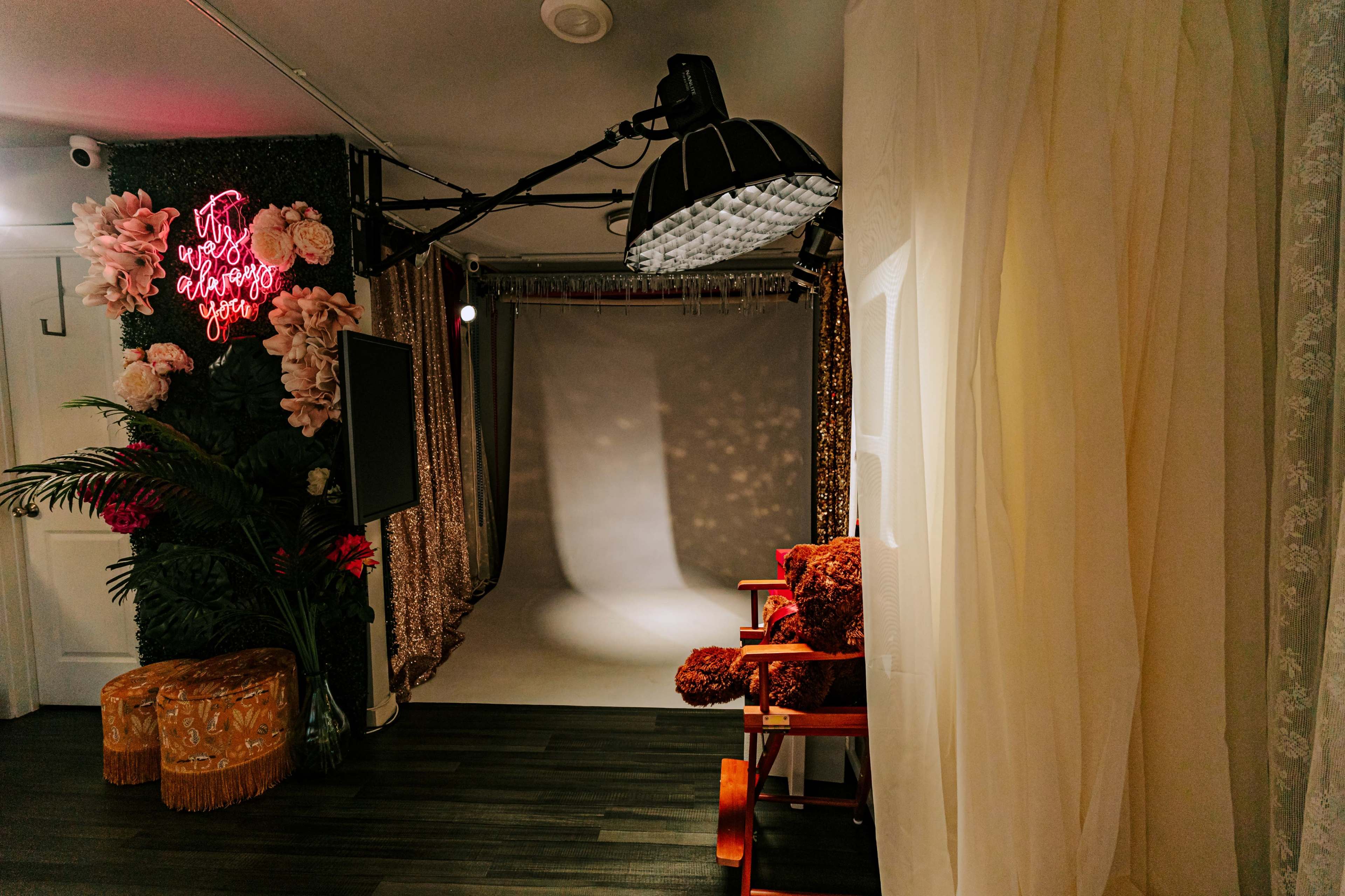 The image shows a photography studio setup featuring a backdrop with decorative elements, bright lighting, and a seating area with red chairs.