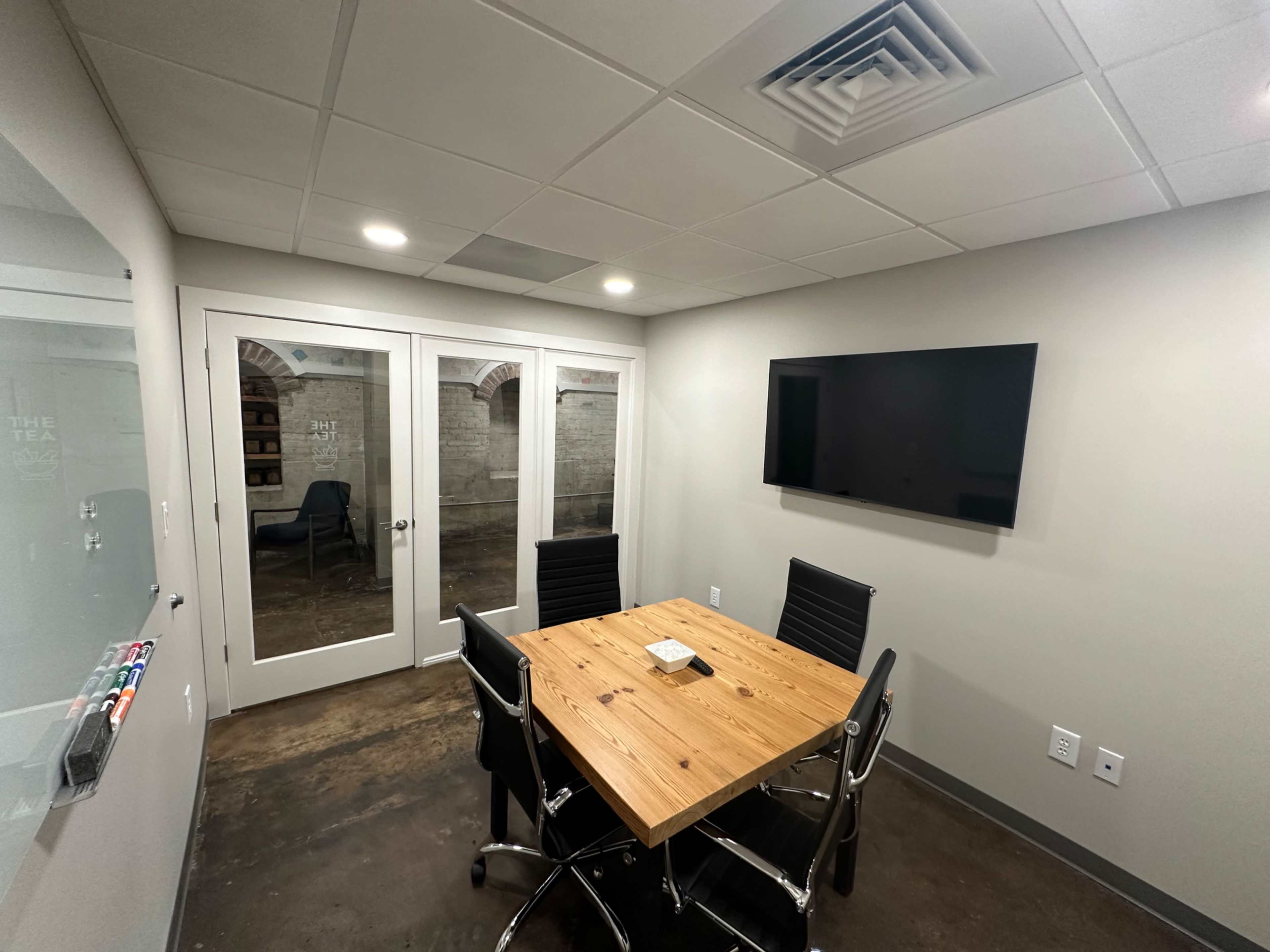 A modern conference room featuring a wooden table with four black chairs, a television mounted on the wall, and glass doors leading to another area.