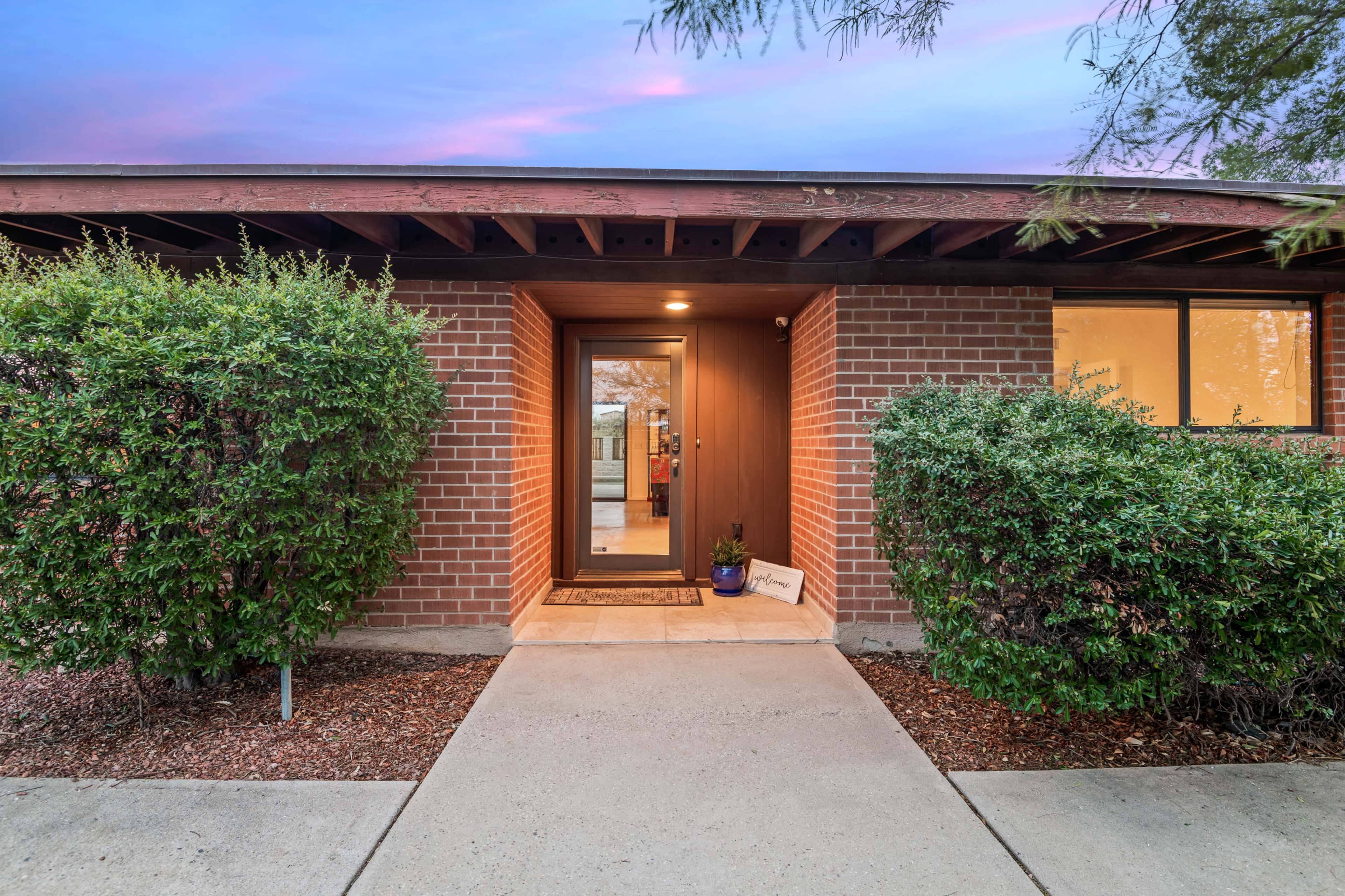 The image shows the entrance of a brick building framed by neatly trimmed hedges and a concrete pathway leading to the door.