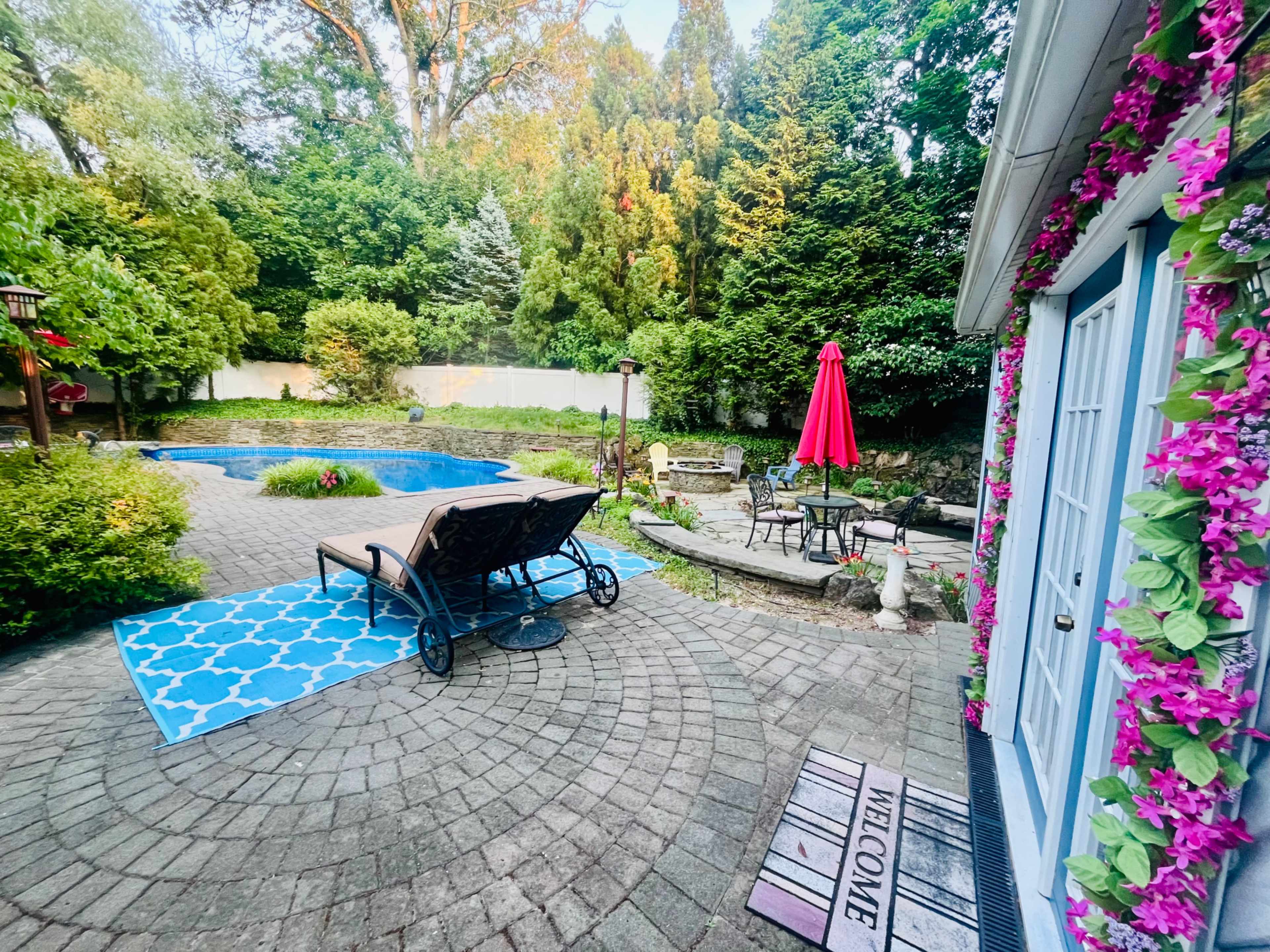 A landscaped backyard with a pool, a lounge chair, a red umbrella, and a patterned rug near a stone pathway.