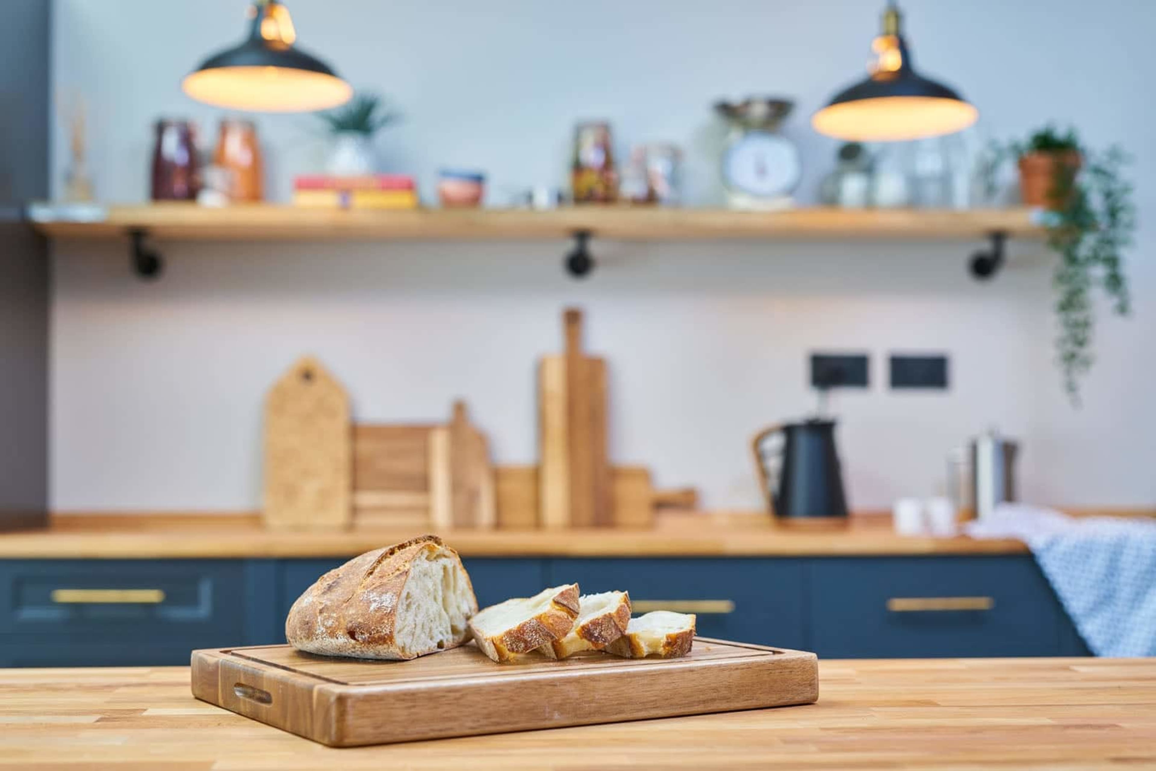 A sliced loaf of bread rests on a wooden cutting board in a kitchen with shelves displaying various items.