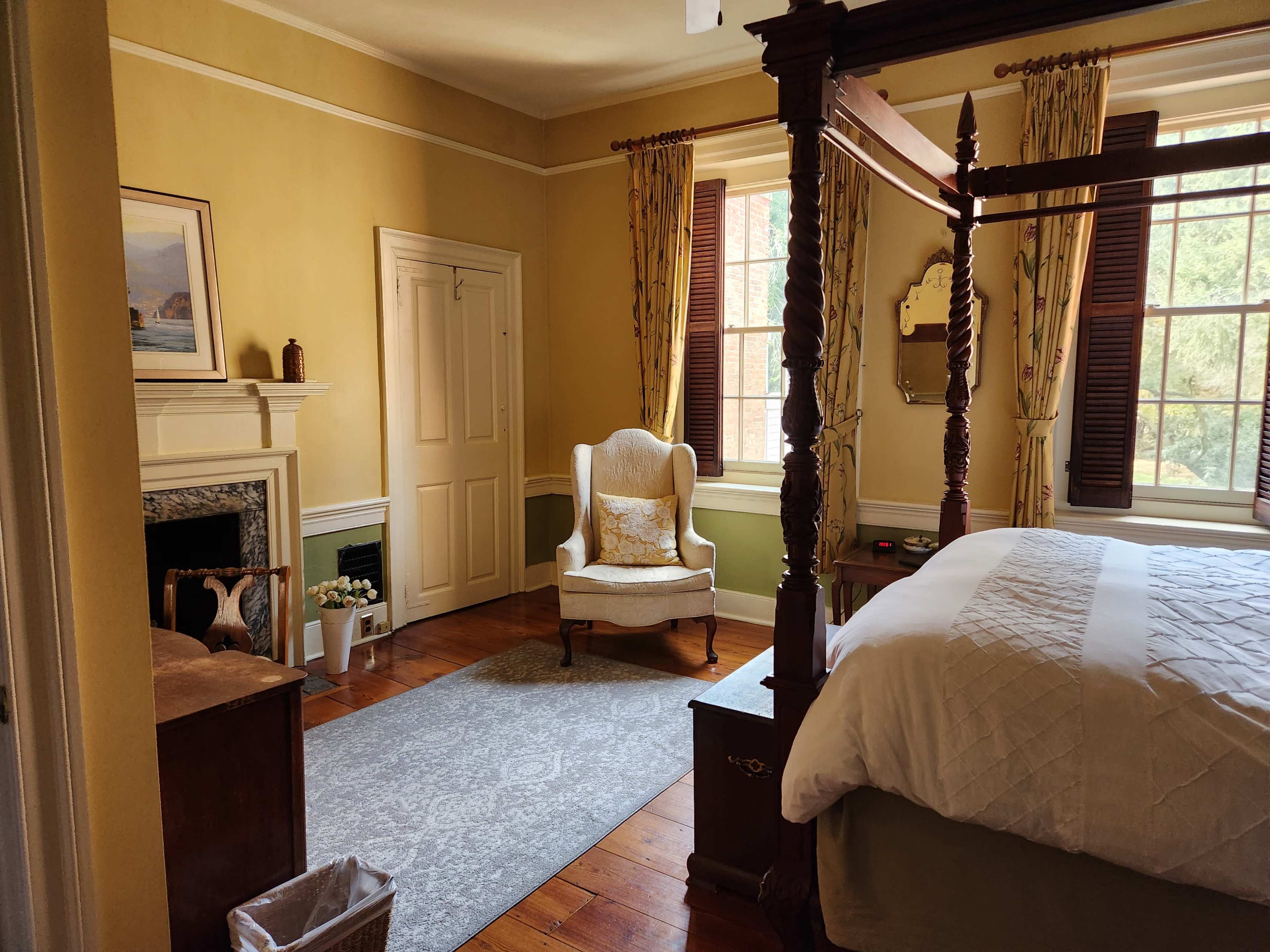 The room features a four-poster bed, a comfortable armchair, a fireplace, and large windows with light yellow walls.