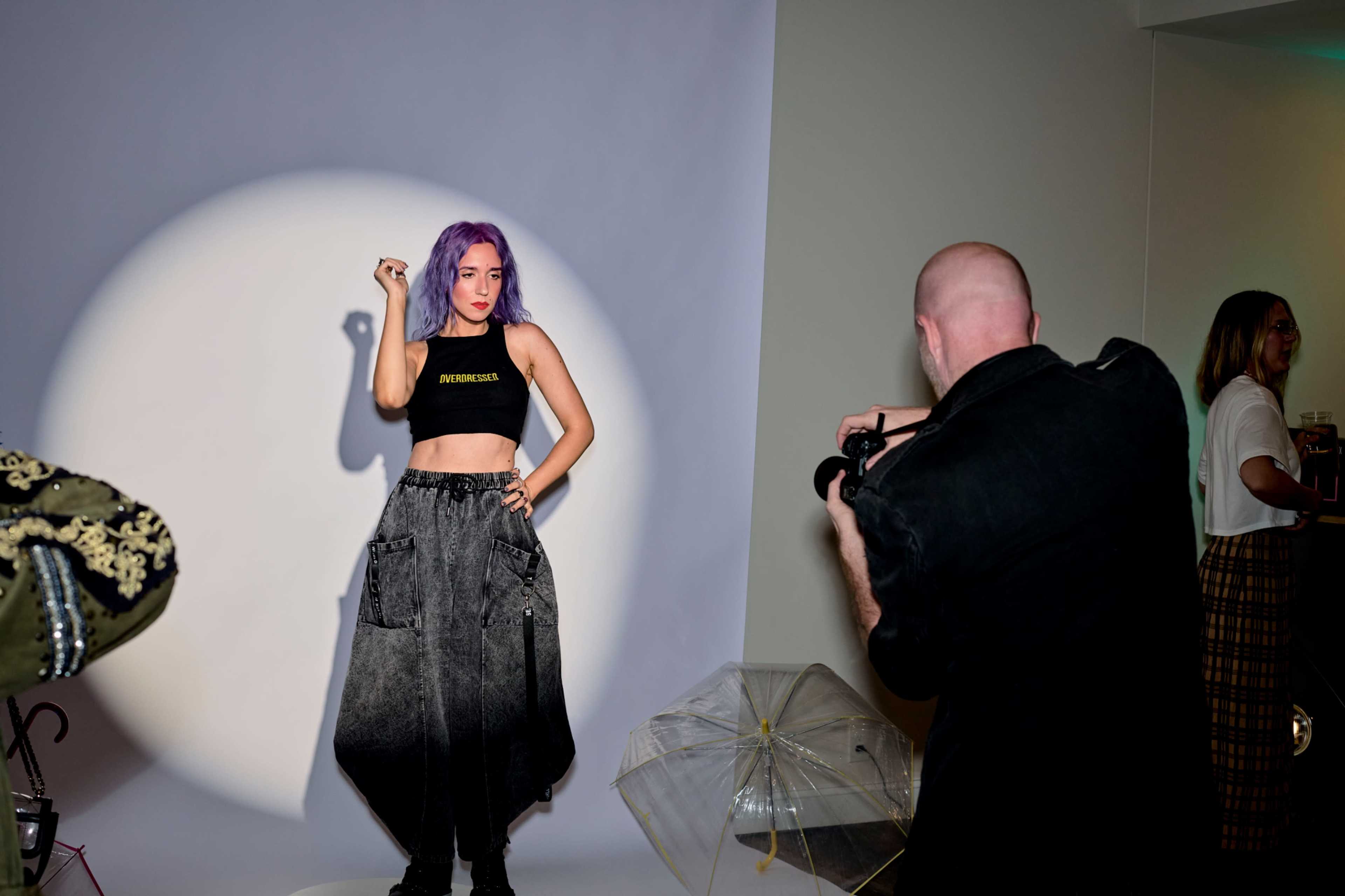 A model with purple hair poses in a black crop top and cargo pants while a photographer captures the scene.