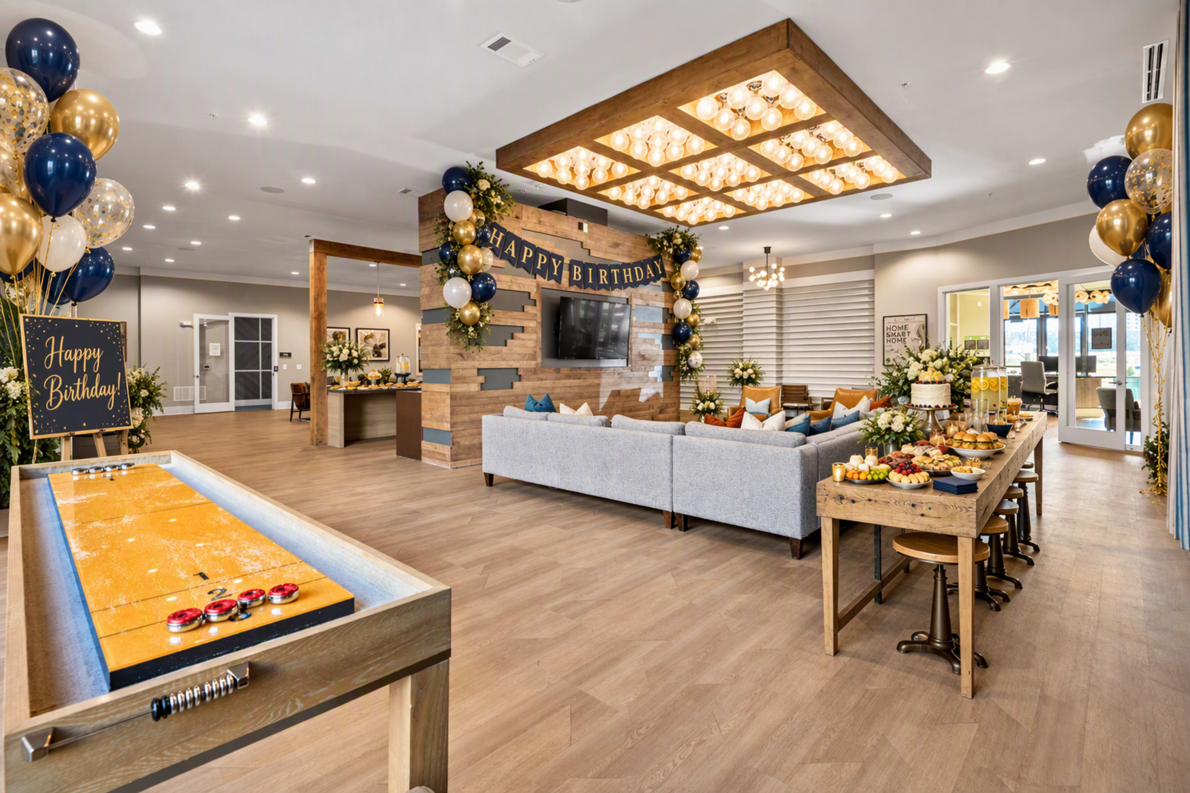 The image shows a decorated party space with comfortable seating, a birthday greeting on the wall, a snack table, and a game area featuring a shuffleboard table.
